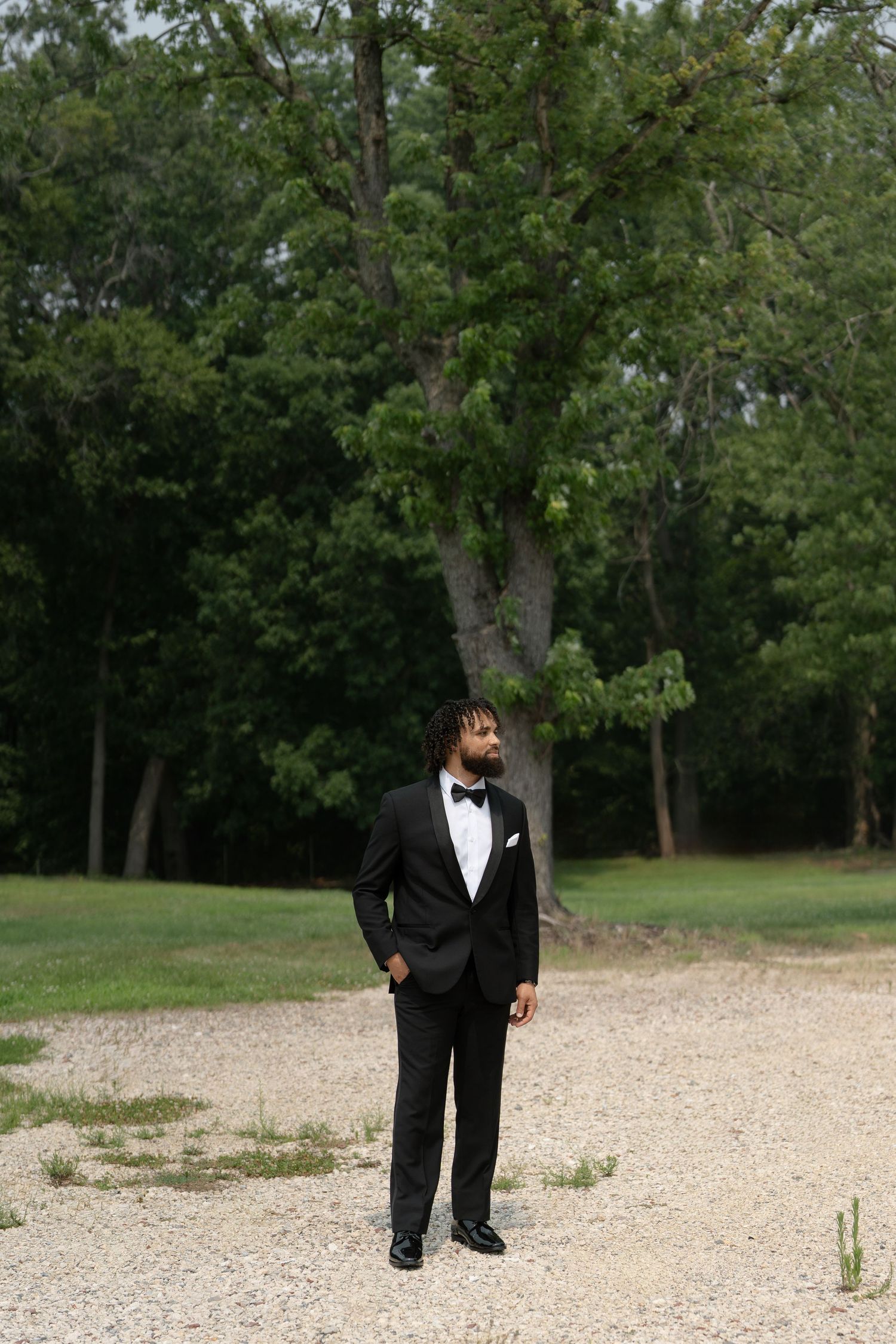 A groom in a black tuxedo with bow tie stands on a gravel path surrounded by lush green trees.