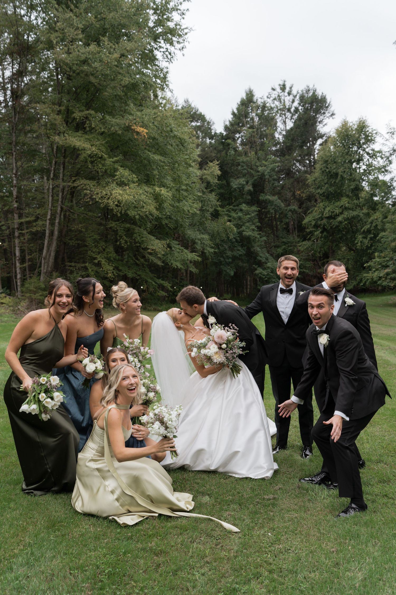 Wedding party laughs and poses playfully on green lawn surrounded by tall forest trees.