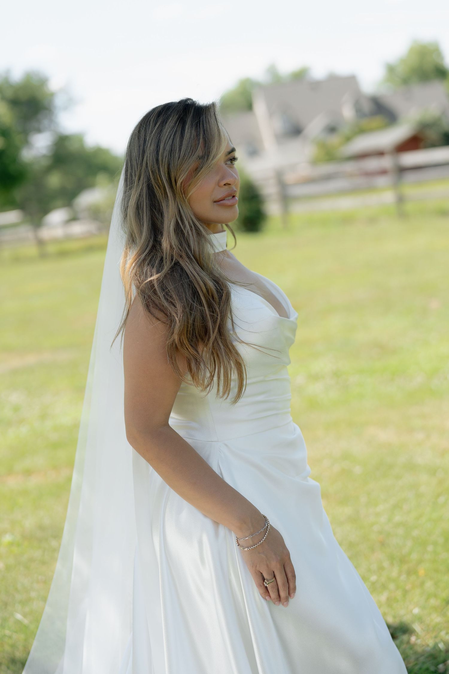 Bride in white strapless gown and veil standing in sunlit green field looking to the side.