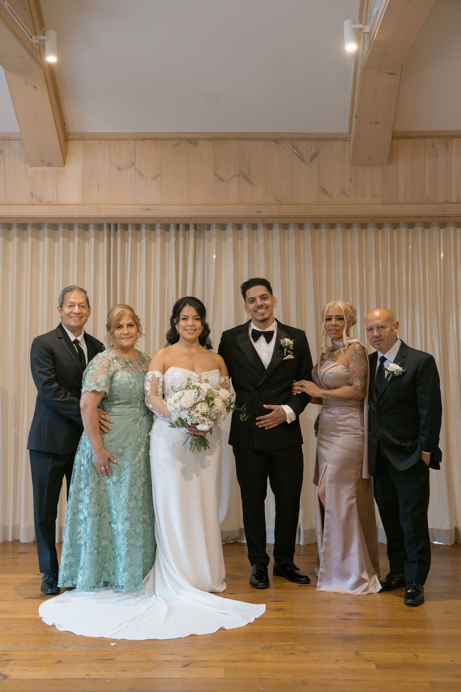 Wedding party portrait featuring bride in white gown, groom in tuxedo, and four family members in formal attire indoors.