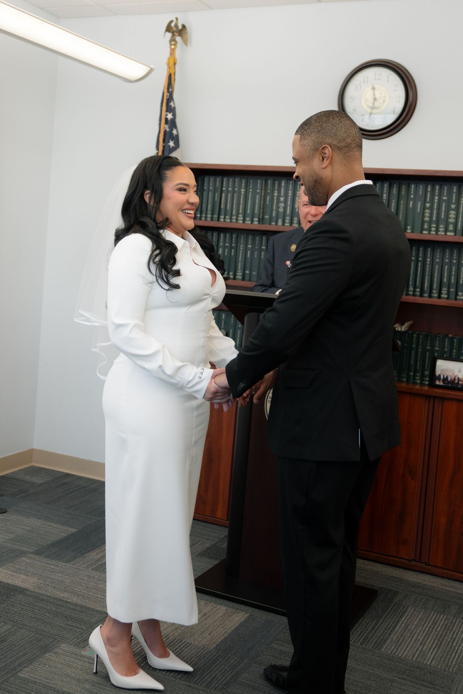A couple in formal attire shares a joyful dance moment in an office setting with bookshelves in the background.