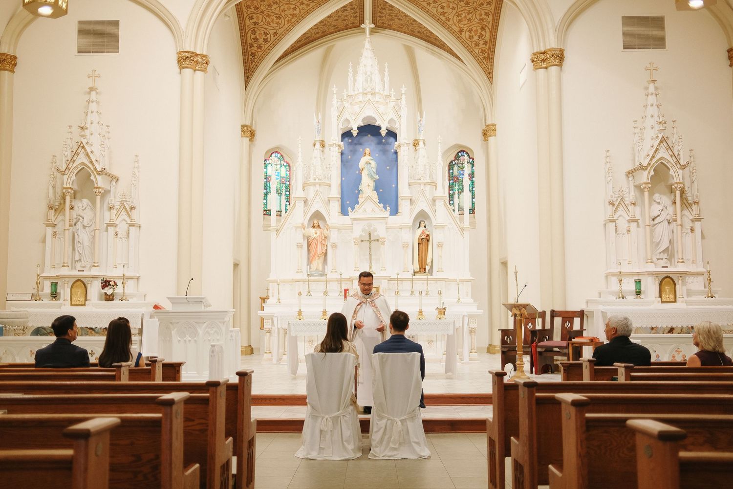 Interior view of a traditional Catholic church sanctuary with white ornate altar, wooden pews and vaulted ceiling details.