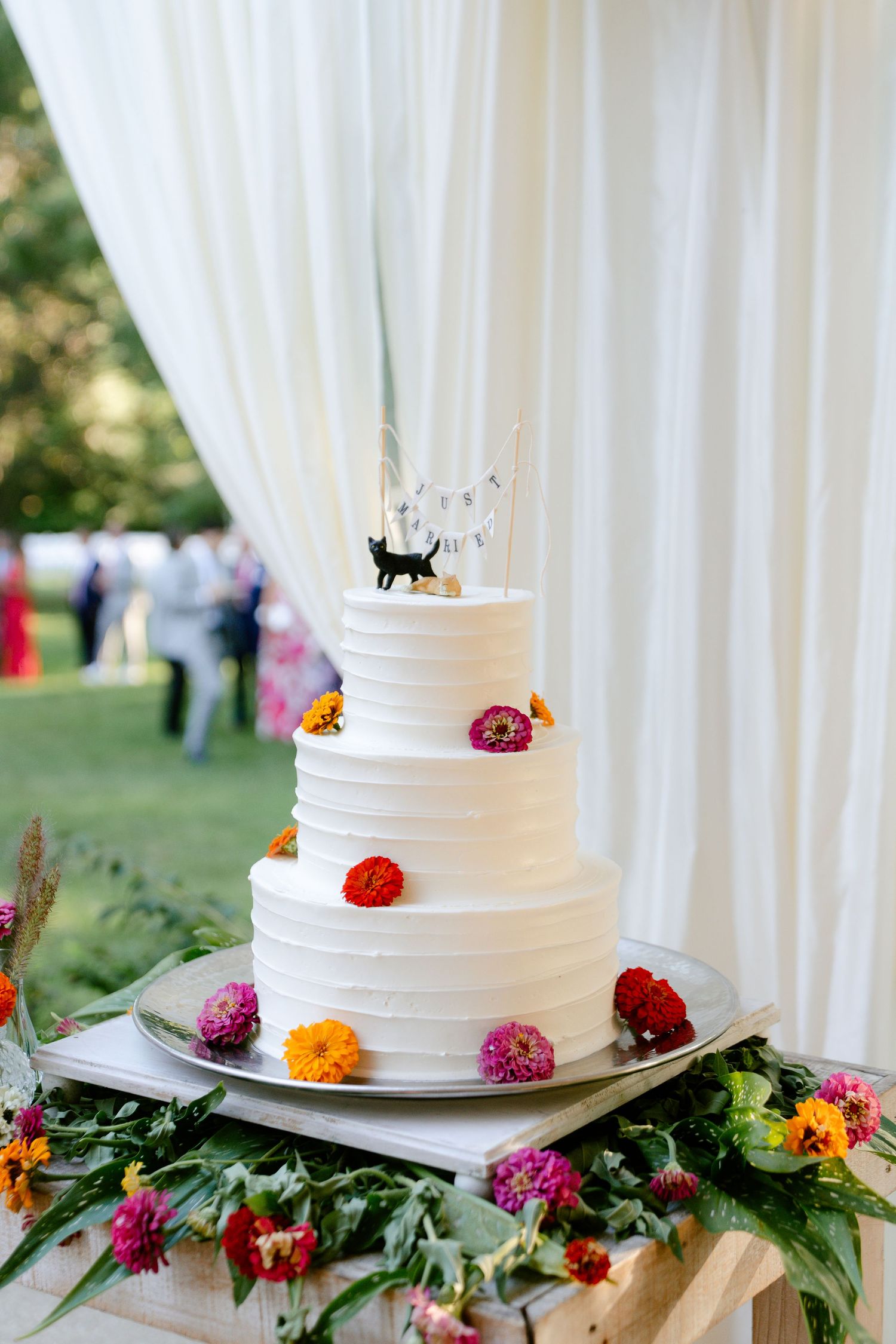 White tiered wedding cake decorated with colorful flowers on a draped table.