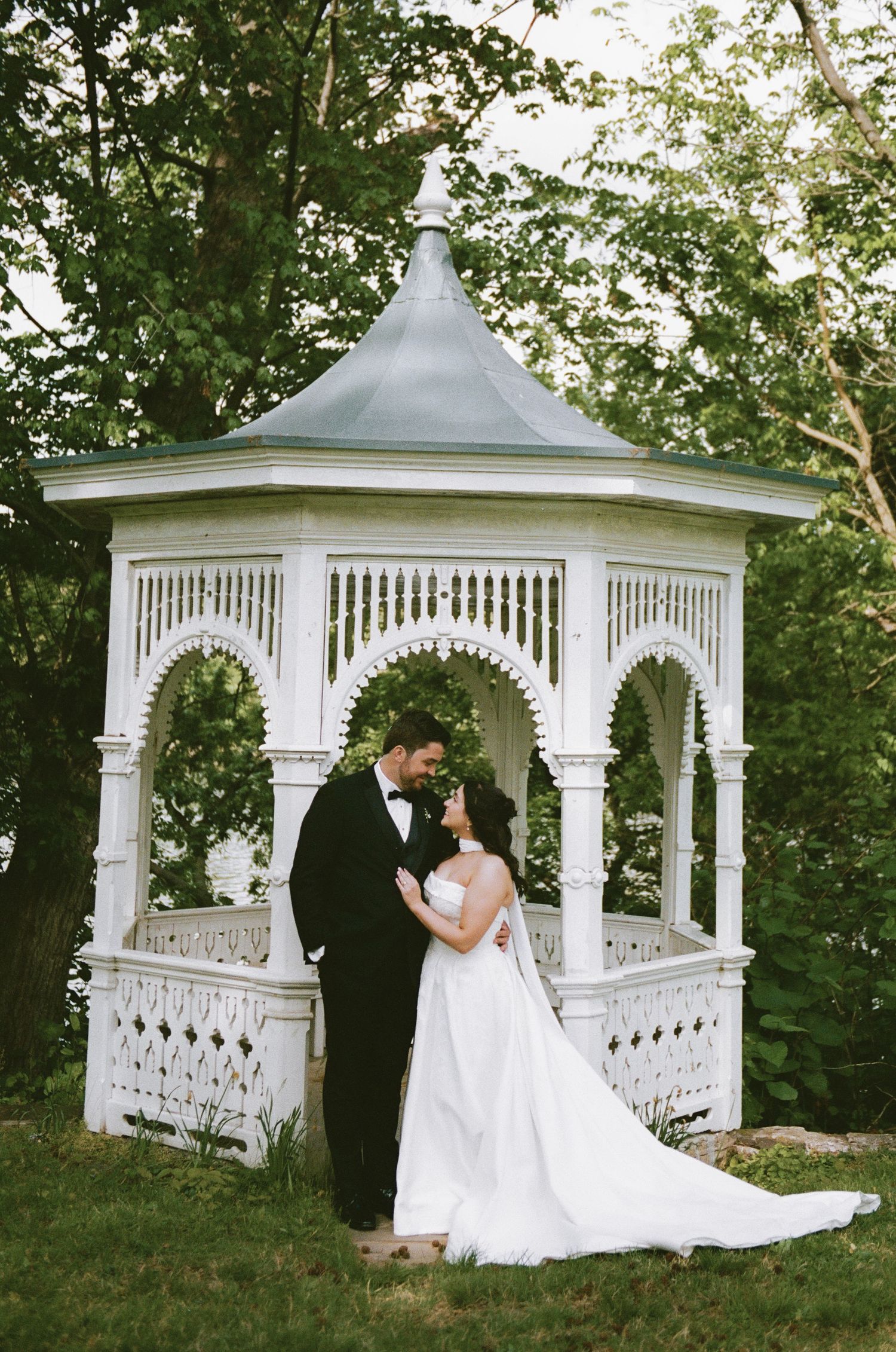 Bride and groom embracing in front of a white Victorian gazebo surrounded by lush green trees on their wedding day.