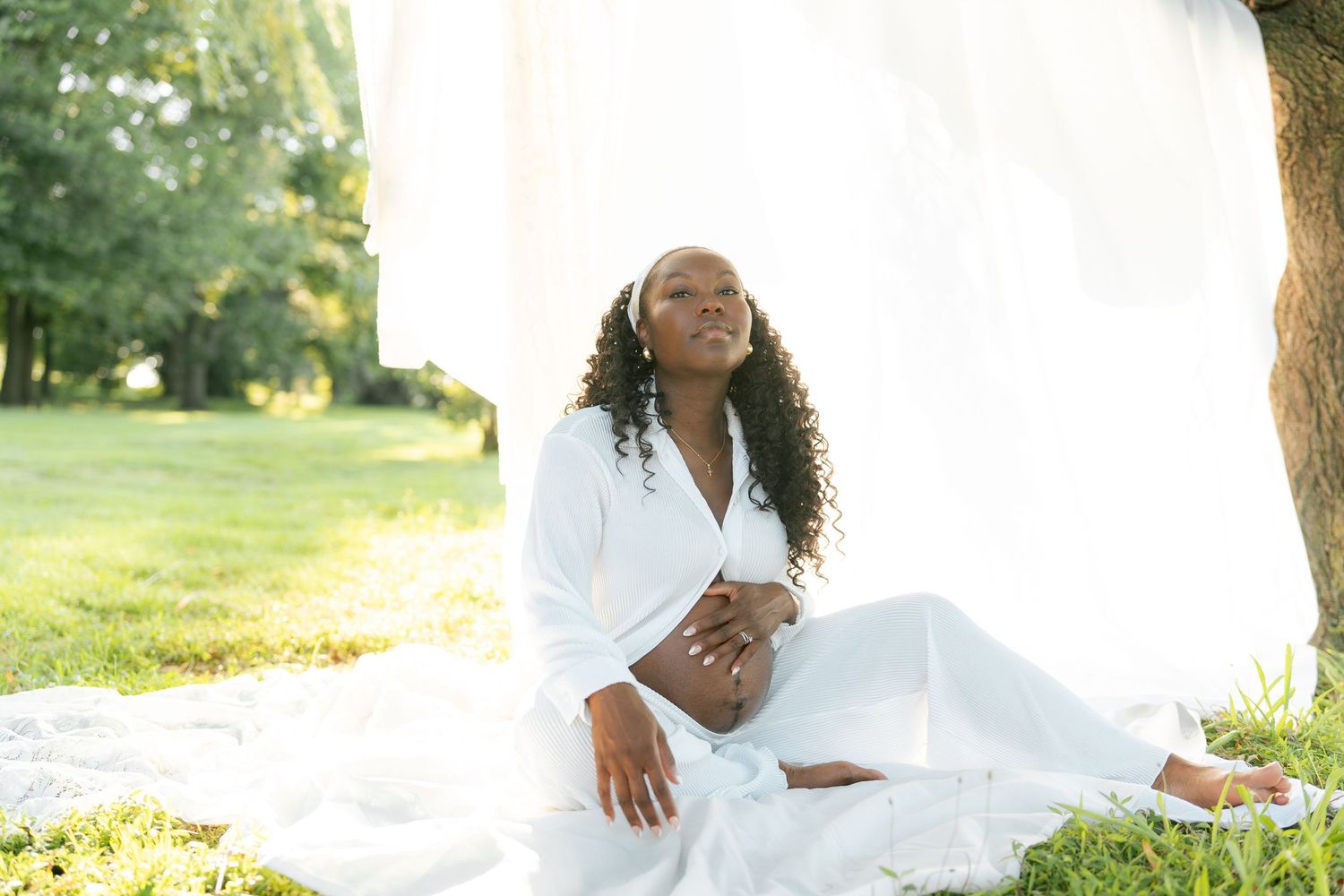 Pregnant Black woman in white outfit sitting on grass outdoors, eyes closed, hands on belly, serene maternity photo.