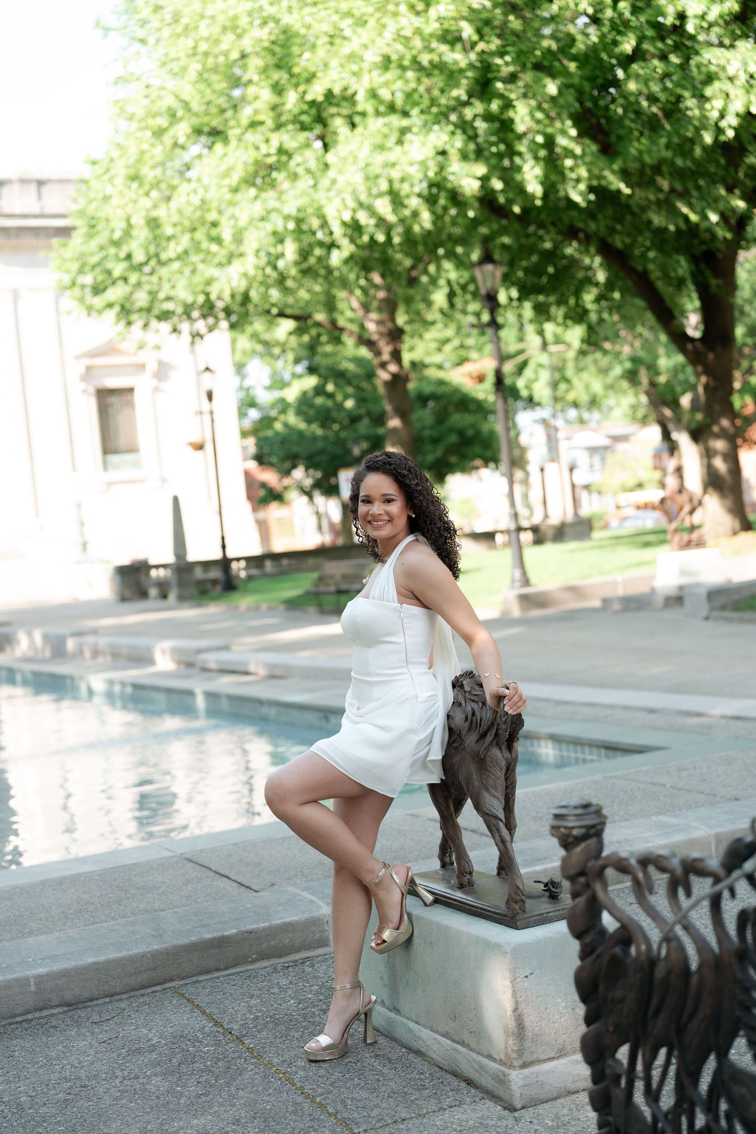 Smiling woman in white dress posing with bronze lion statue near fountain in sunny outdoor plaza.