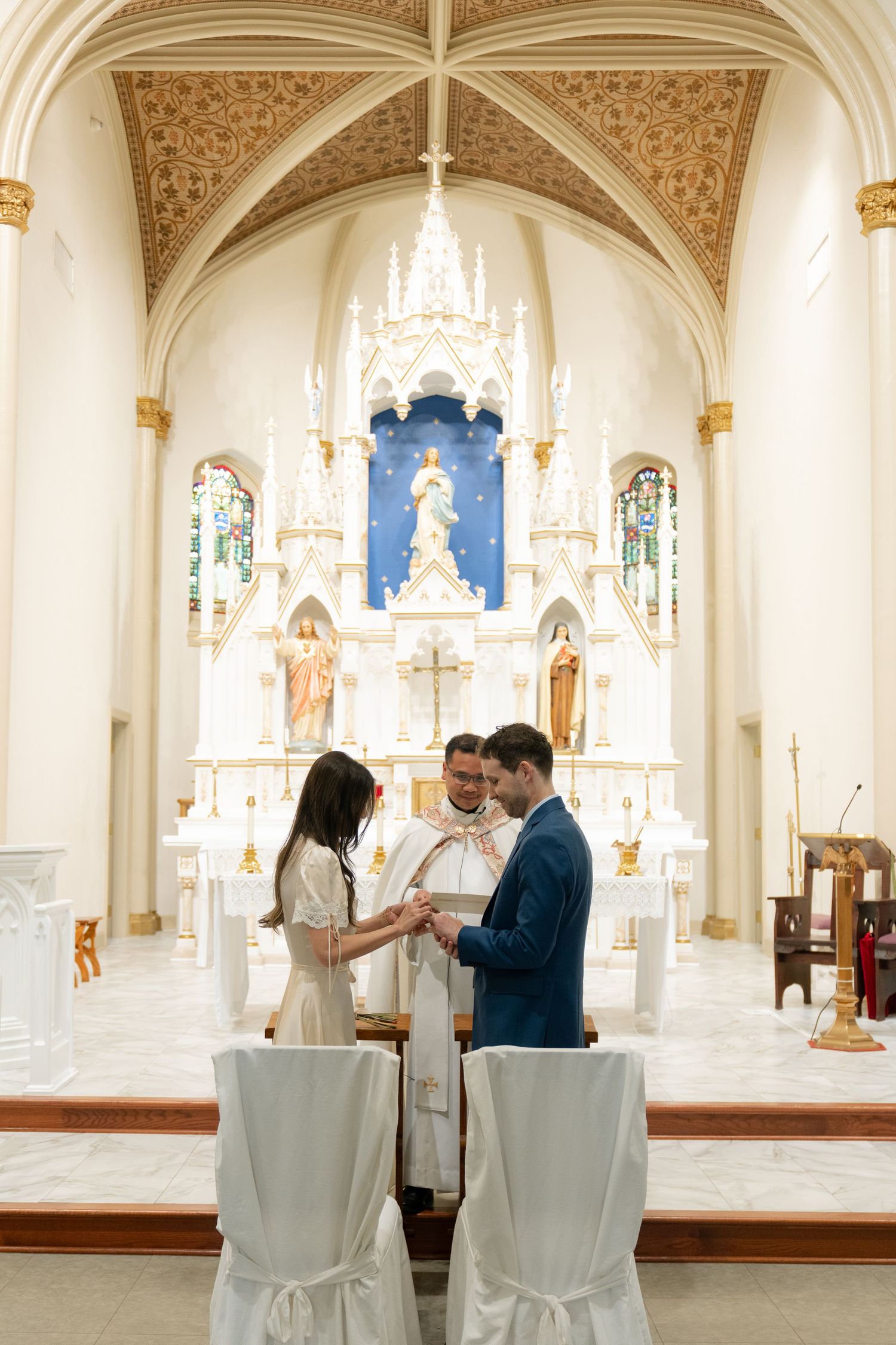 A wedding ceremony takes place in an ornate Catholic church with vaulted ceilings and white and gold decor.