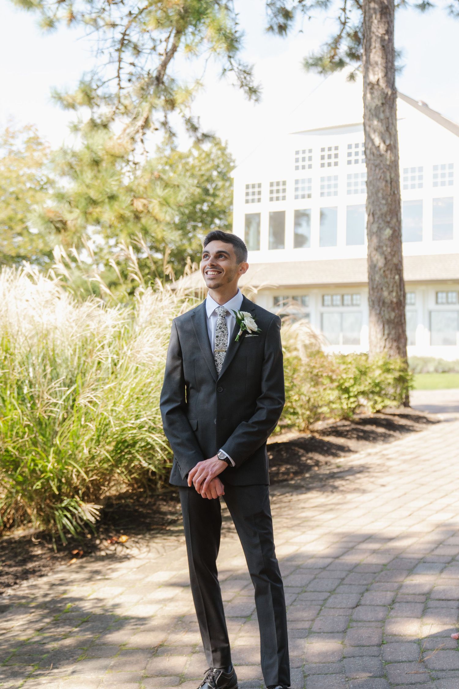 A groom in a dark suit stands on a stone pathway surrounded by lush greenery and pine trees on a sunny day.