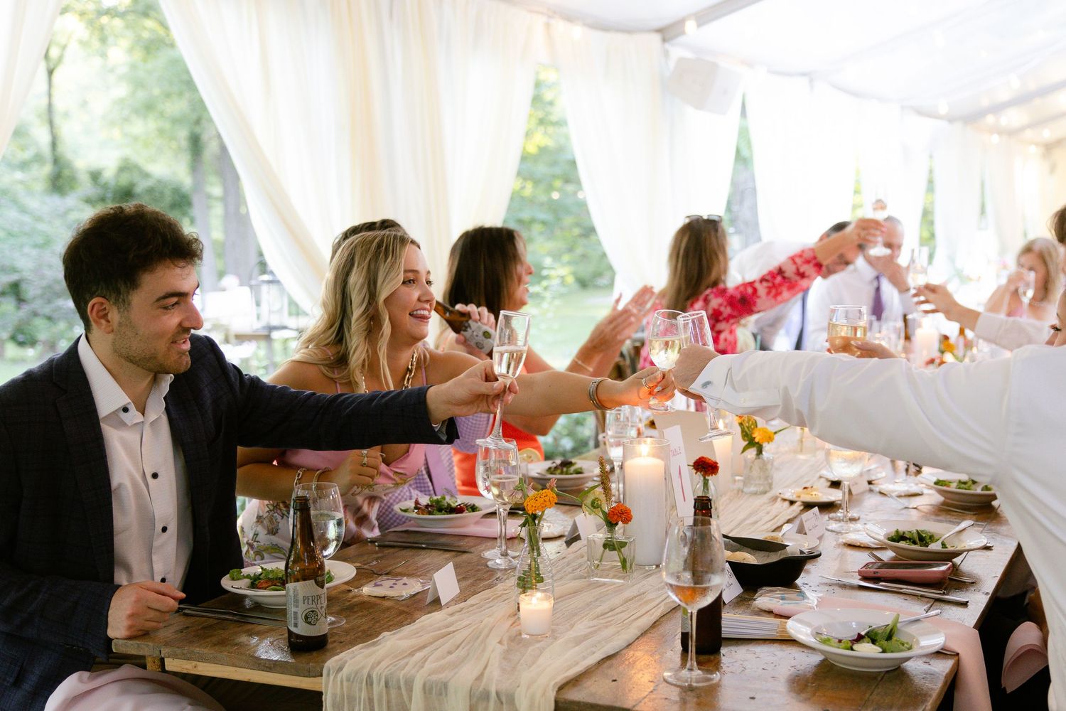 Wedding reception guests raise their glasses in a toast at a decorated table under a tent.