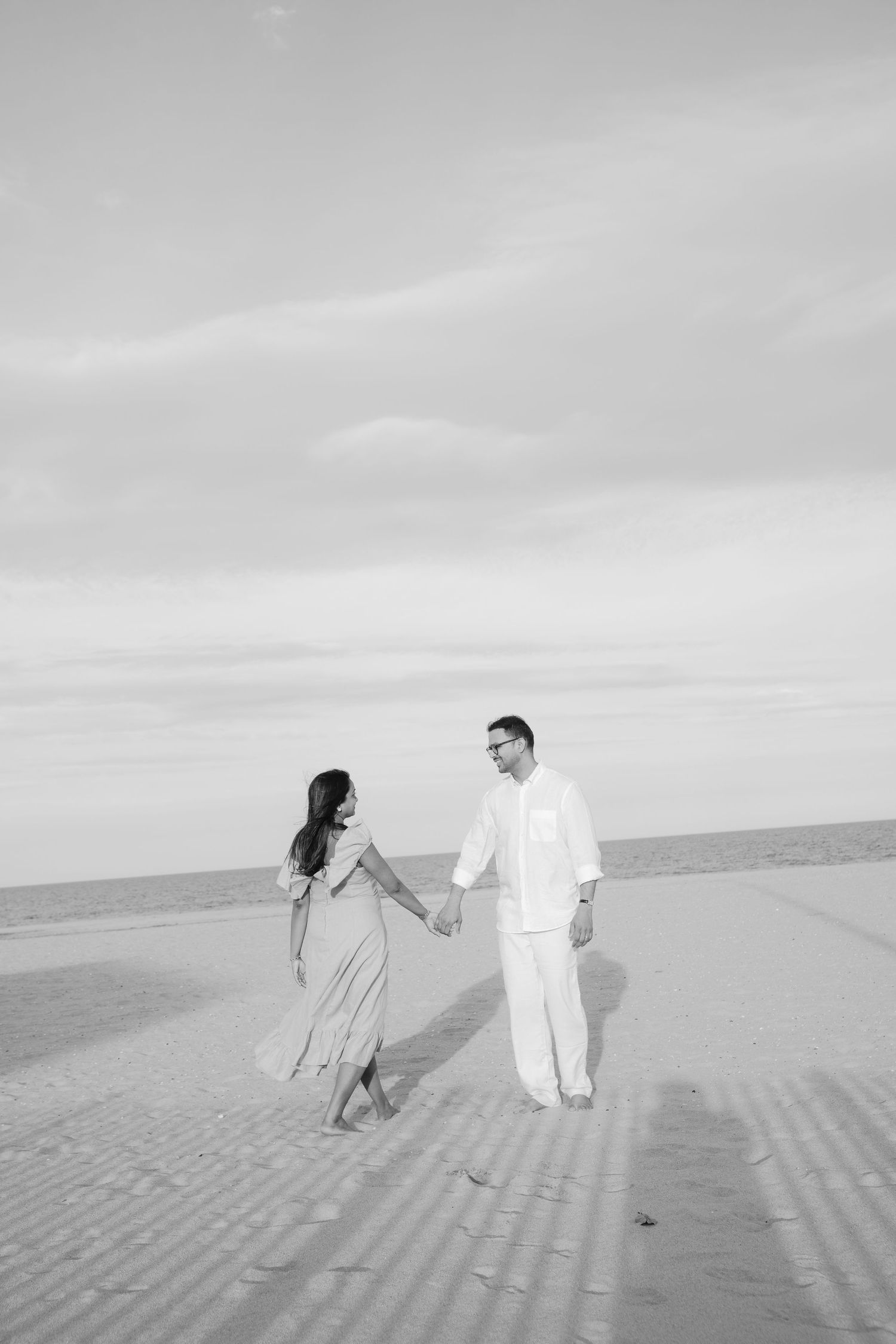 Couple walking hand in hand along beach shoreline in ethereal black and white photography.