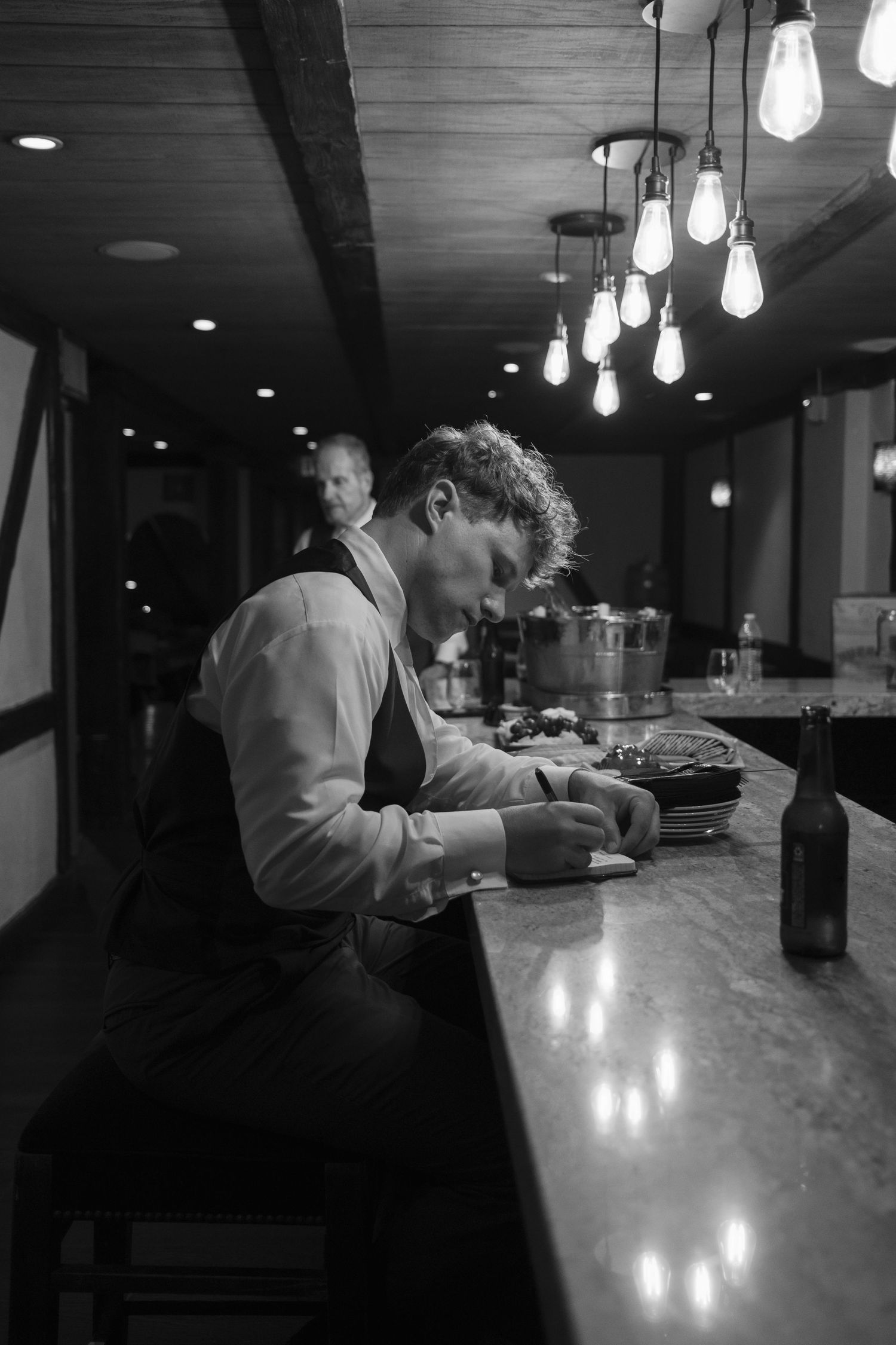 Black and white photo of a bartender working at a dimly lit bar with vintage Edison bulb lighting overhead.