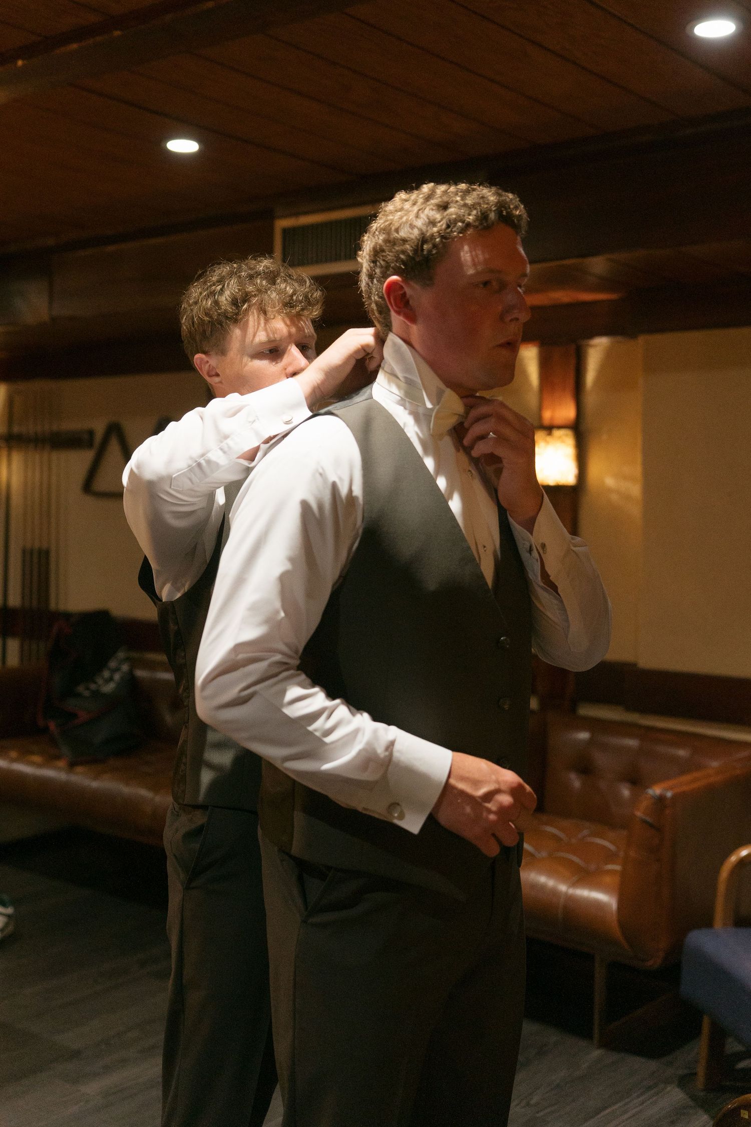 Groomsman helping groom adjust his tie while wearing a gray vest and white dress shirt before a wedding.