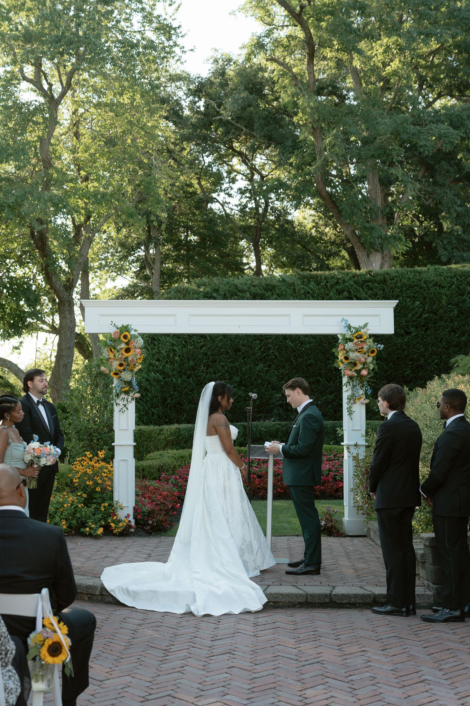 Bride and groom exchange vows under a white floral arch in an outdoor garden wedding ceremony.