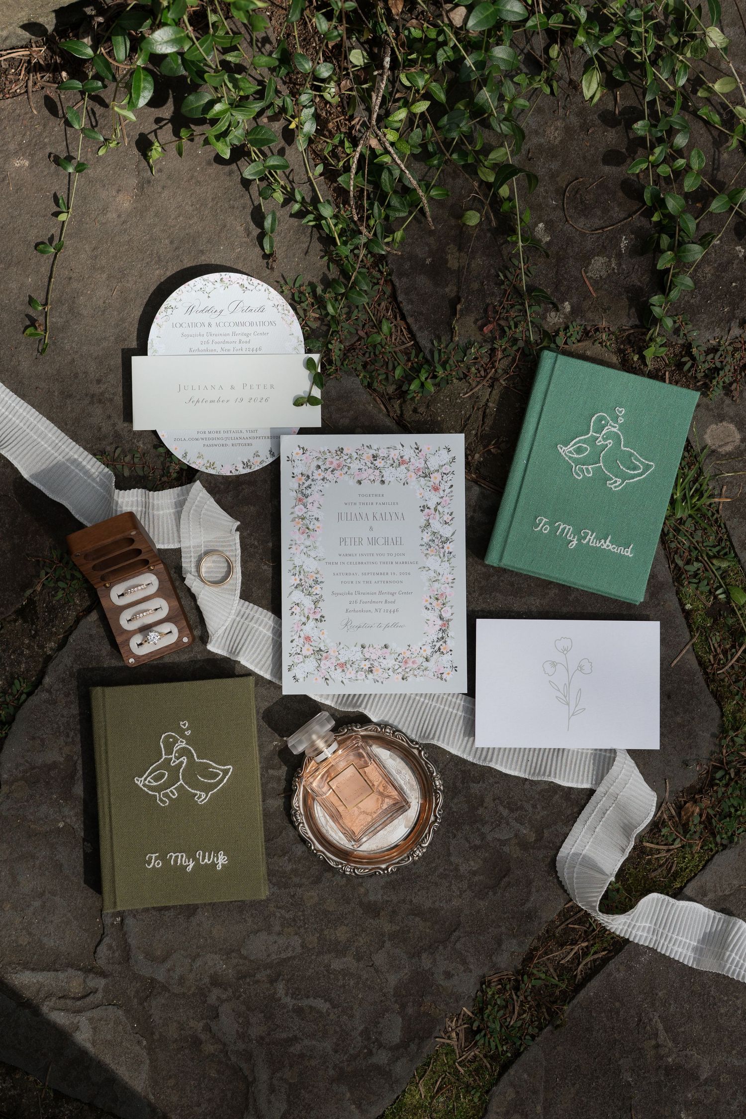 Elegant wedding stationery flat lay featuring floral invitations, vow books, perfume, and ribbon on stone.