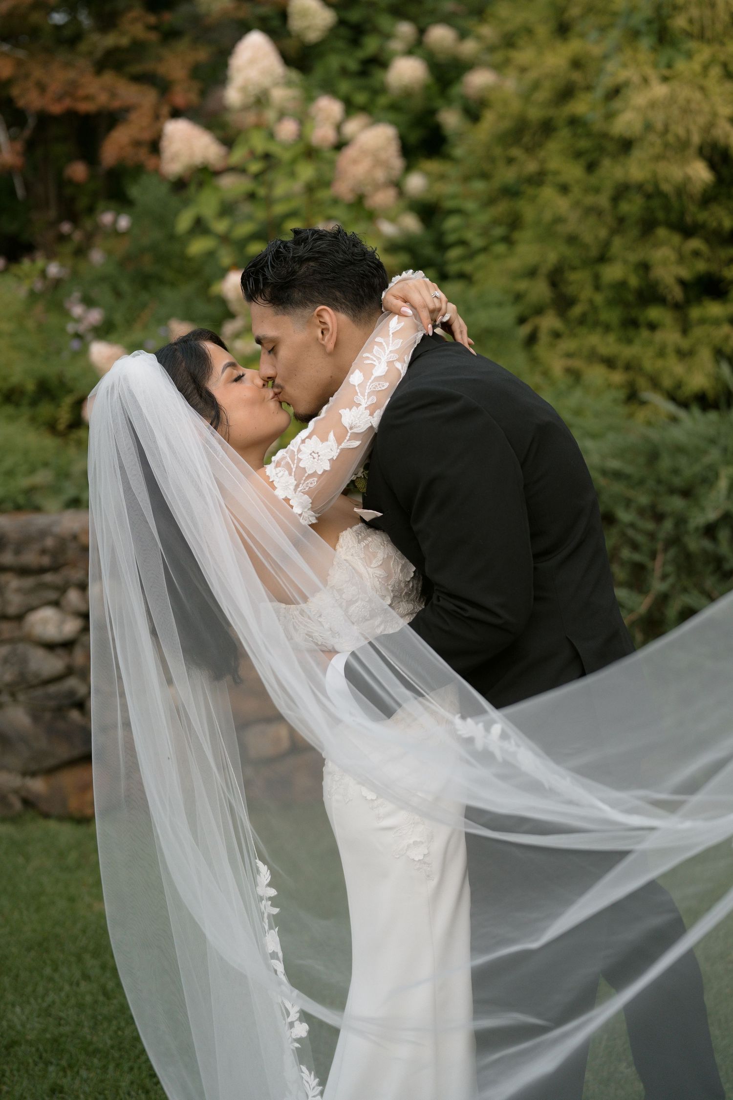 Groom dips bride for a romantic kiss outdoors, her lace wedding dress and flowing veil billowing in the breeze.