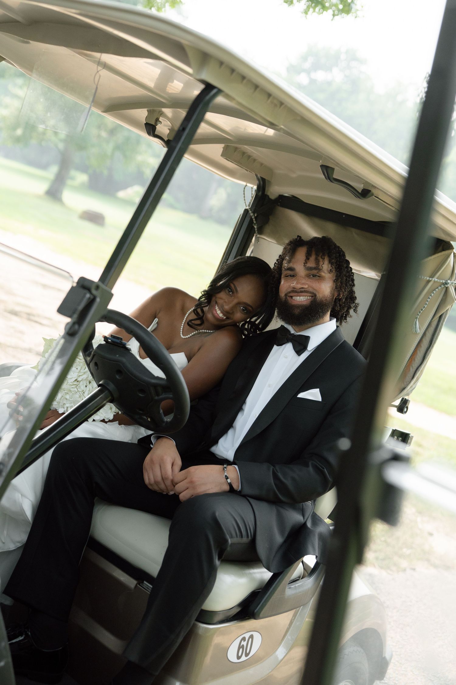 Smiling bride and groom in formal attire sitting closely together in a golf cart at an outdoor venue.