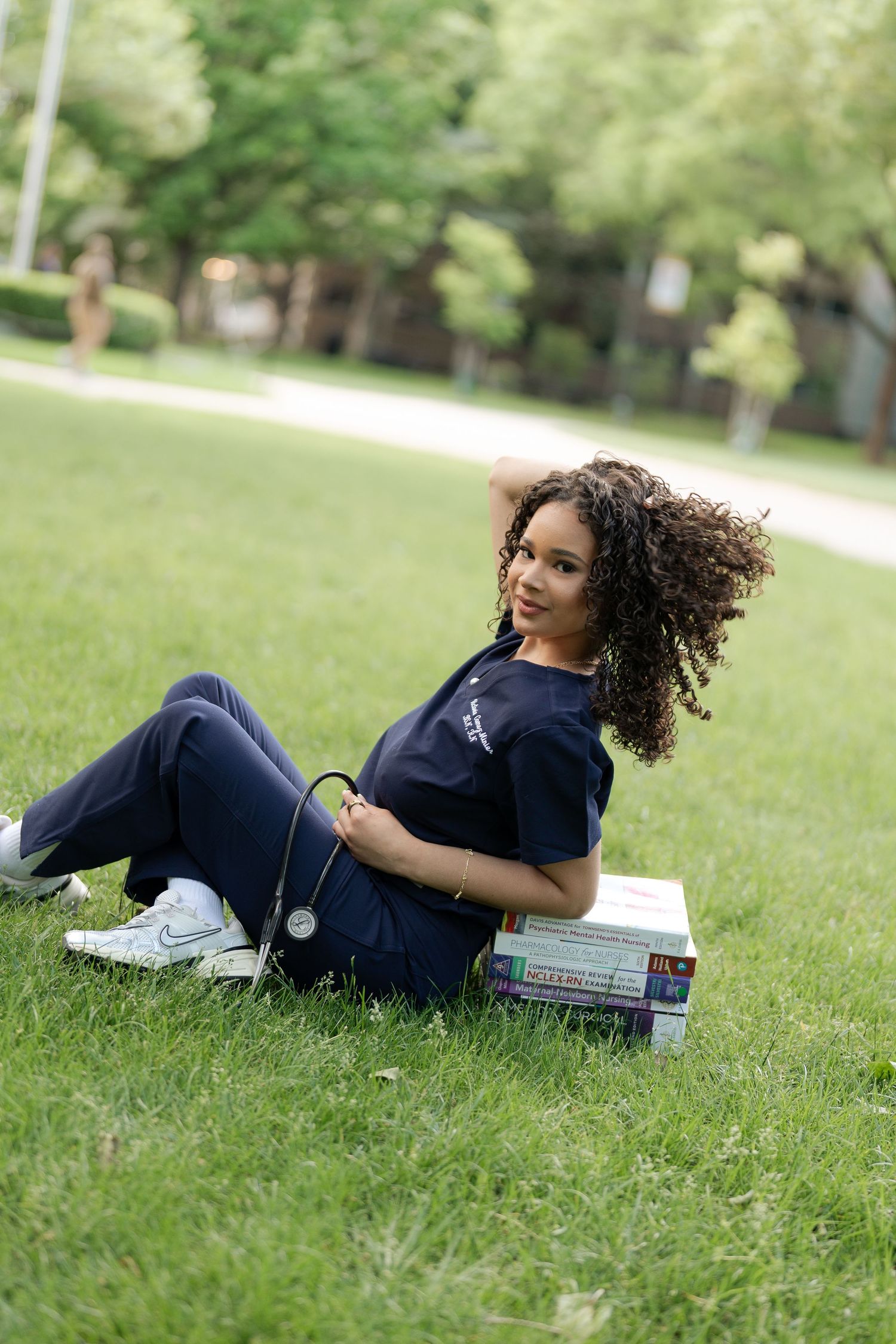 Nursing student in scrubs relaxes on grass with medical textbooks and stethoscope.