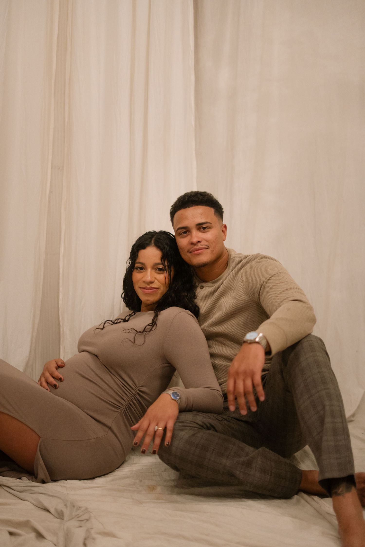 A smiling couple poses together on the floor against white curtains, woman in tan bodysuit showing her baby bump.