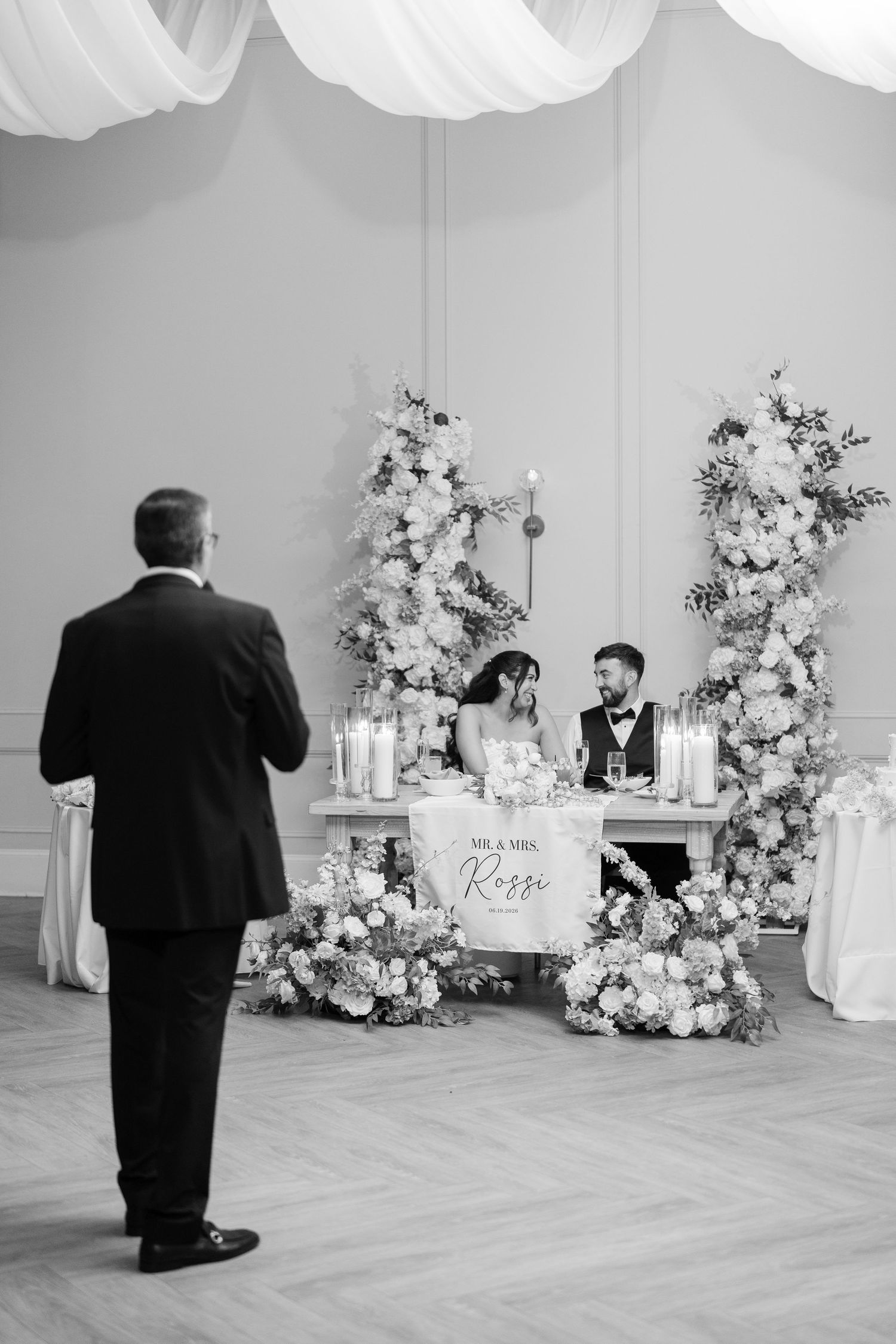 Black and white photo of Mr. and Mrs. Rossi seated at sweetheart table during wedding reception speech.