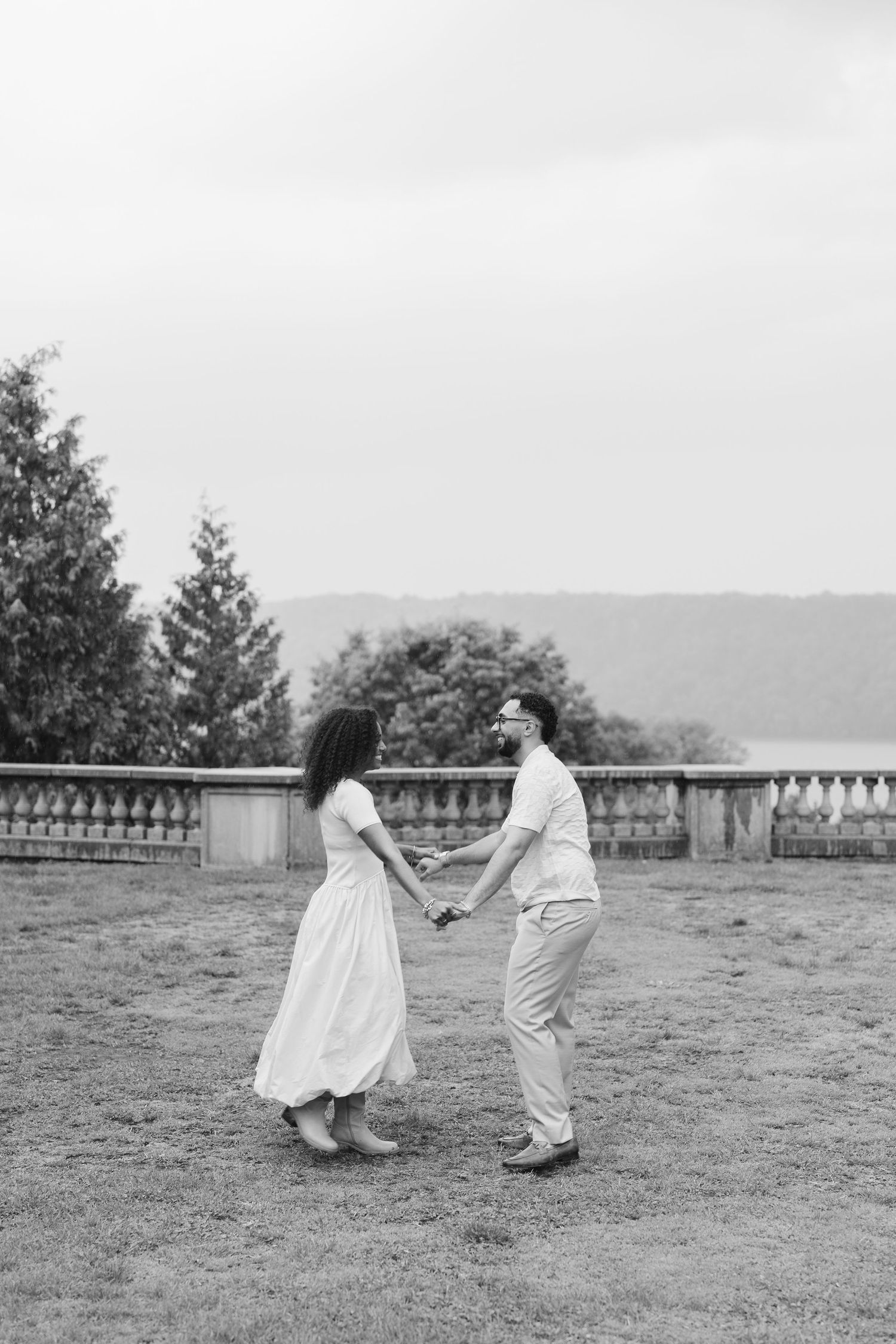 Black and white photo of a couple holding hands and dancing on a garden terrace with trees and water in the background.