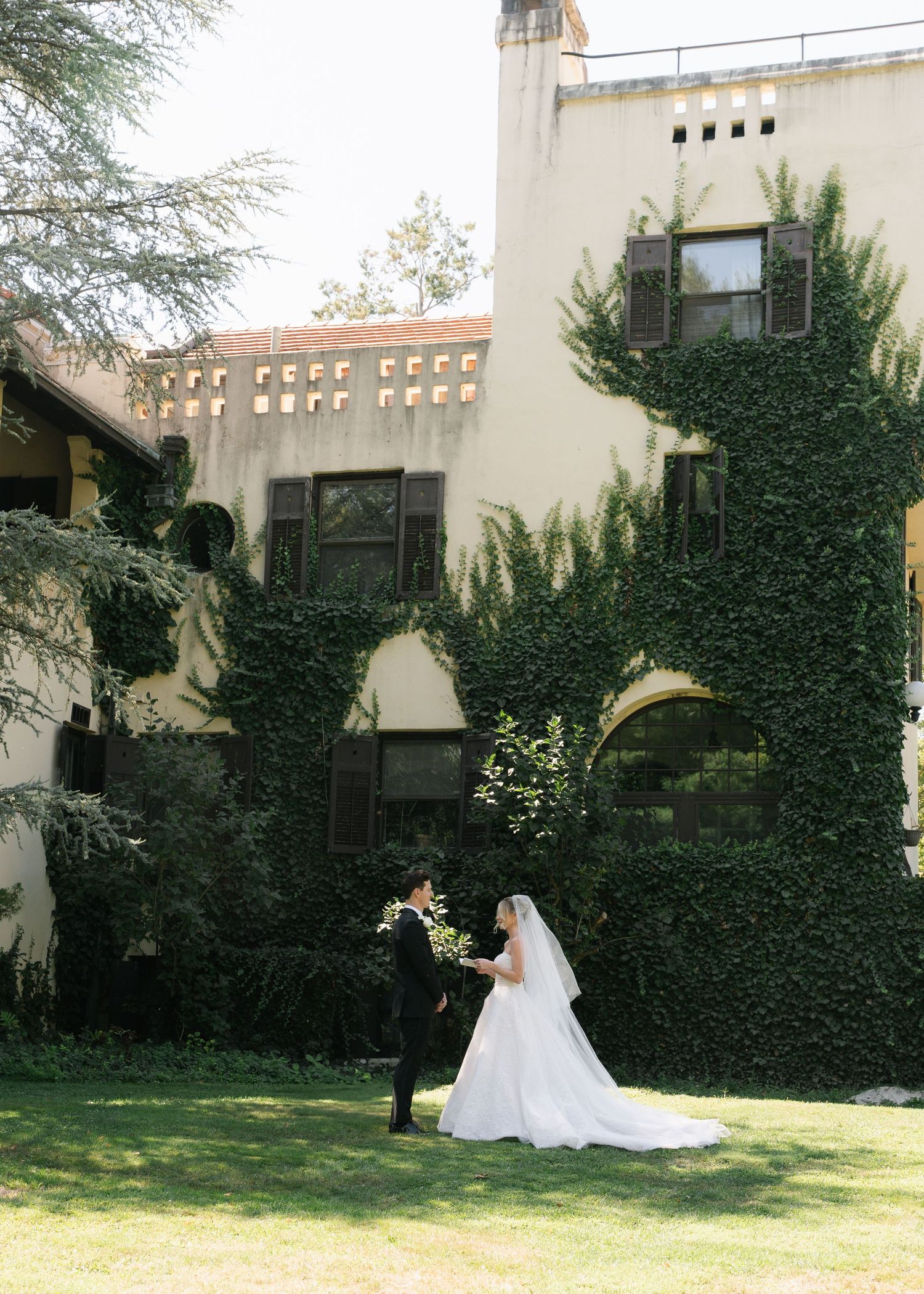 A bride and groom share a romantic moment on a lush green lawn beside an ivy-covered estate wedding venue.