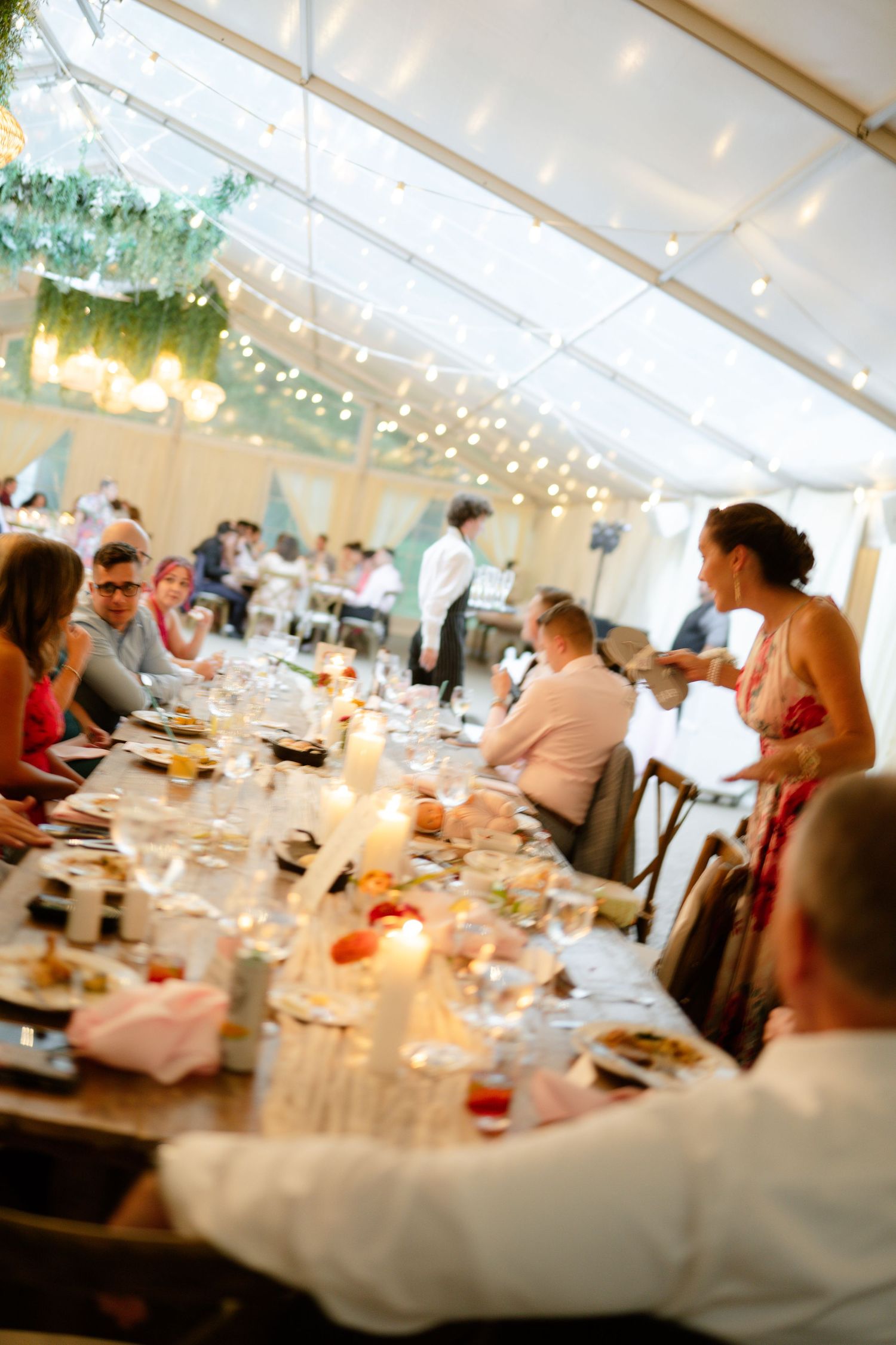 Wedding reception under white tent with tables set for dinner and string lights overhead.
