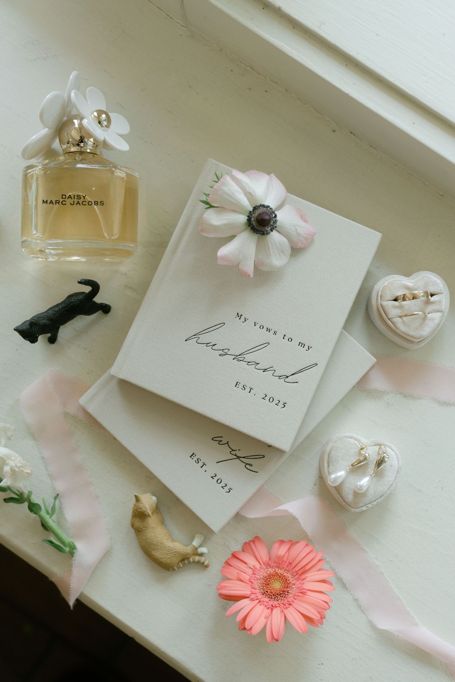 Wedding details including invitation cards, perfume bottles, and delicate pink floral decorations arranged on white surface.