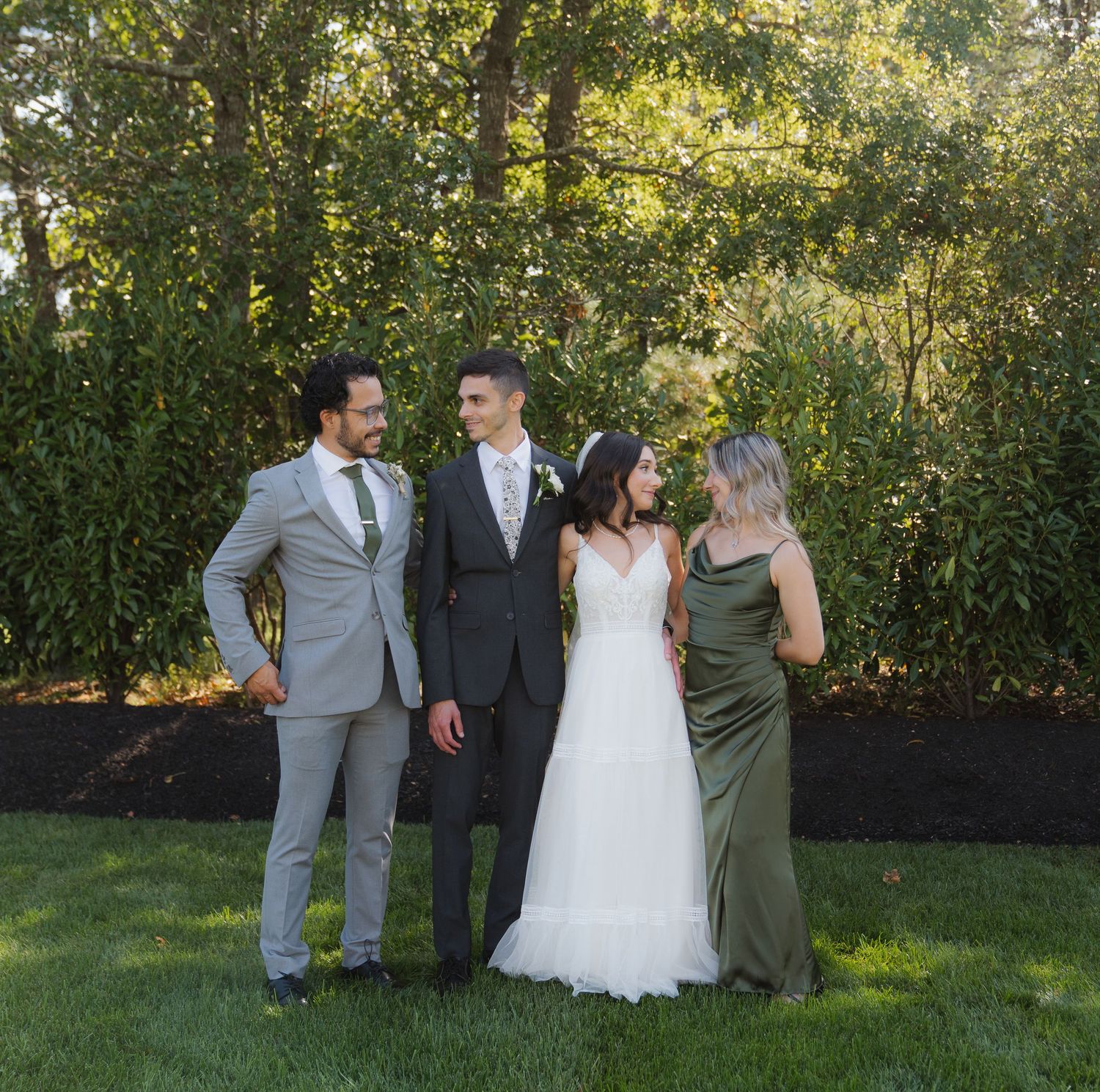 Wedding party in gray and navy suits with white dress pose together on a sunny lawn with green foliage background.