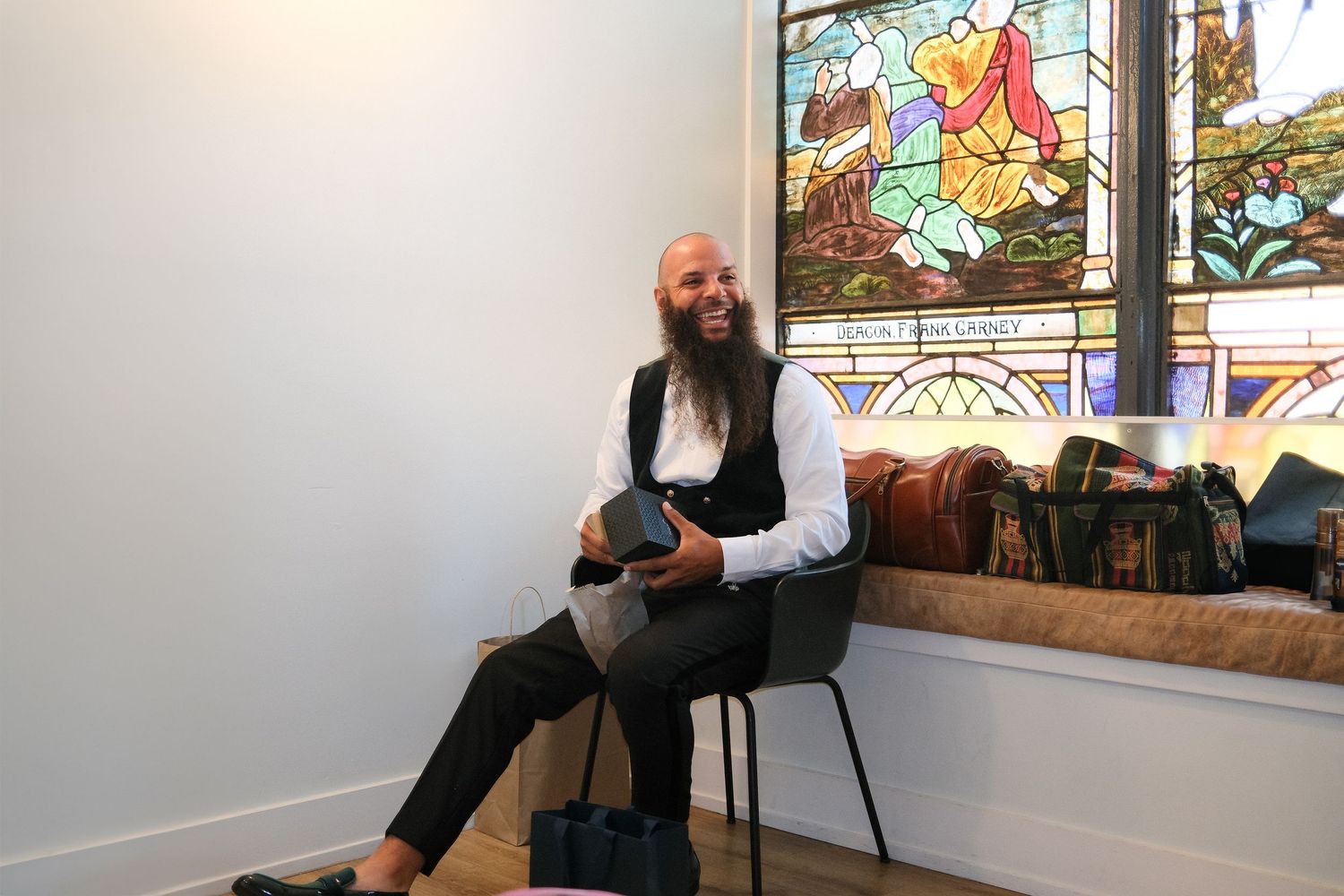 A person in formal attire sits in a chair by a stained glass window in an elegant room setting.