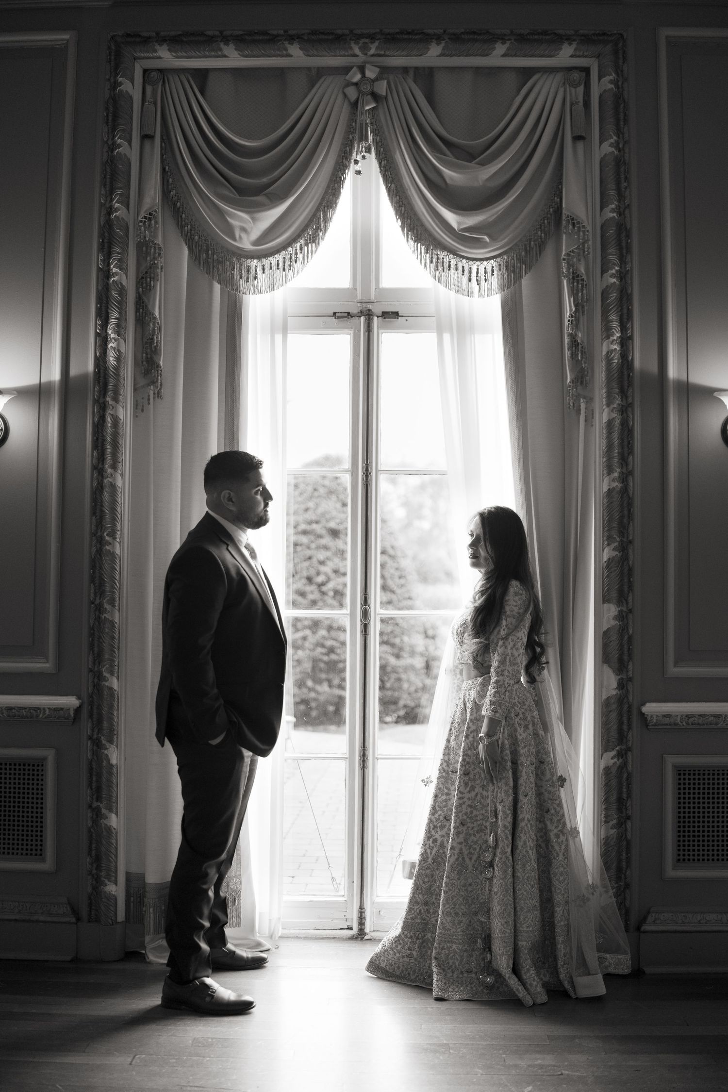 Dramatic black and white photo of a couple by a window in an elegant room with flowing curtains.