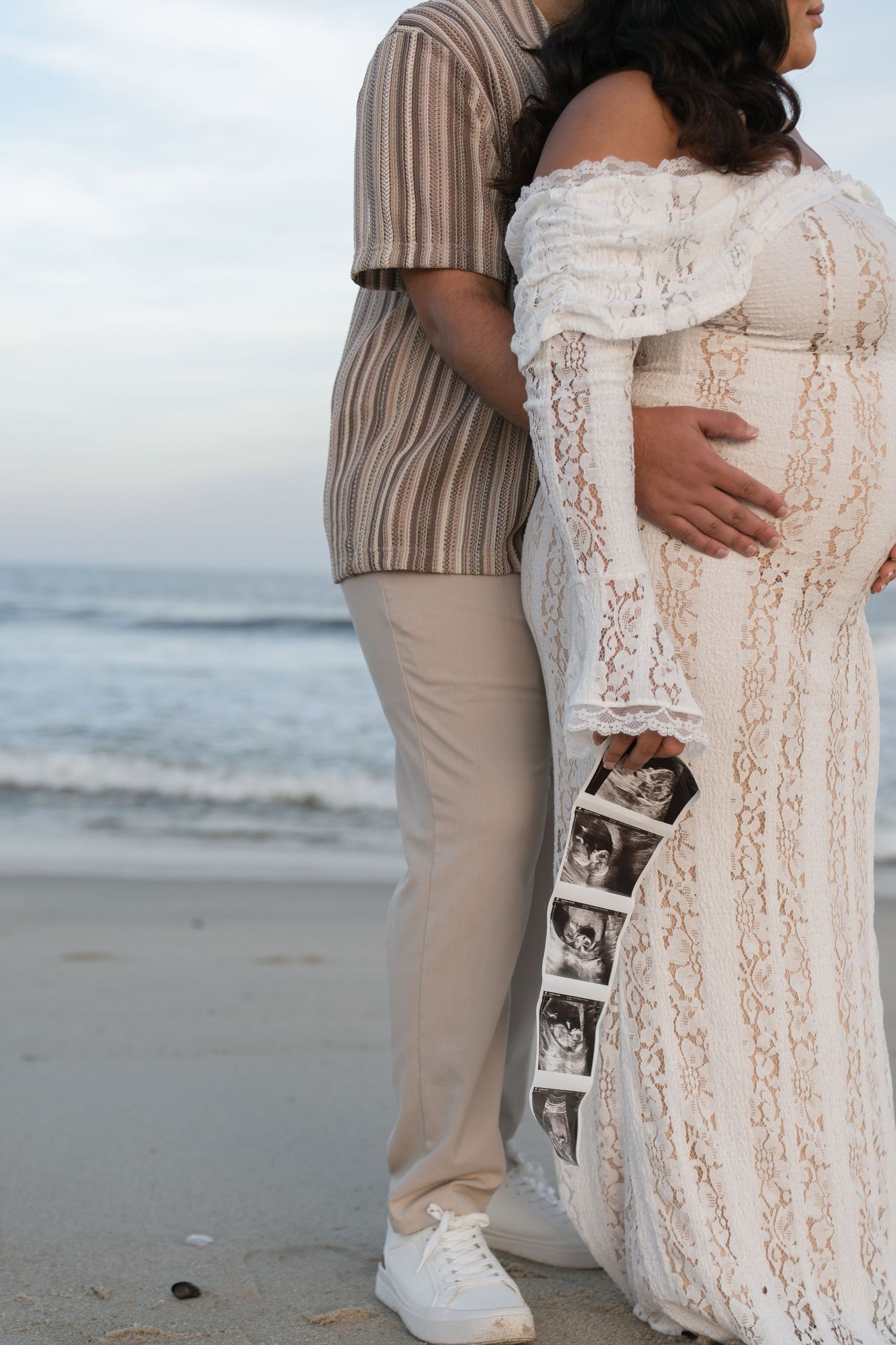 Pregnant couple embracing on beach, woman in white lace maternity gown holding ultrasound photos by ocean.