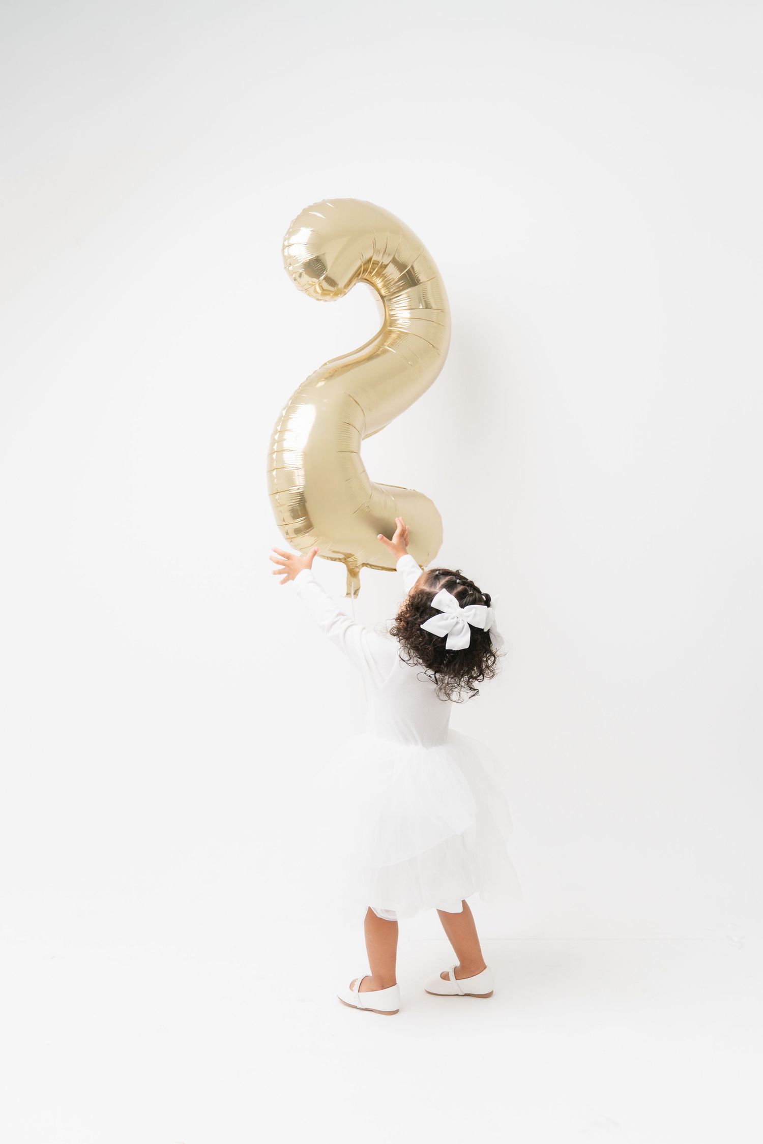 A child in a white dress holds a large gold number 2 balloon against a white background.
