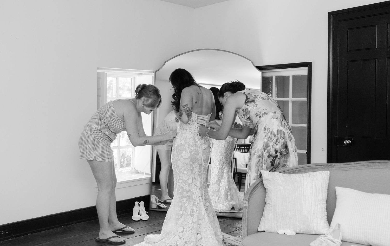 A black and white photograph showing people gathered around a mirror in a room helping with formal attire preparations.