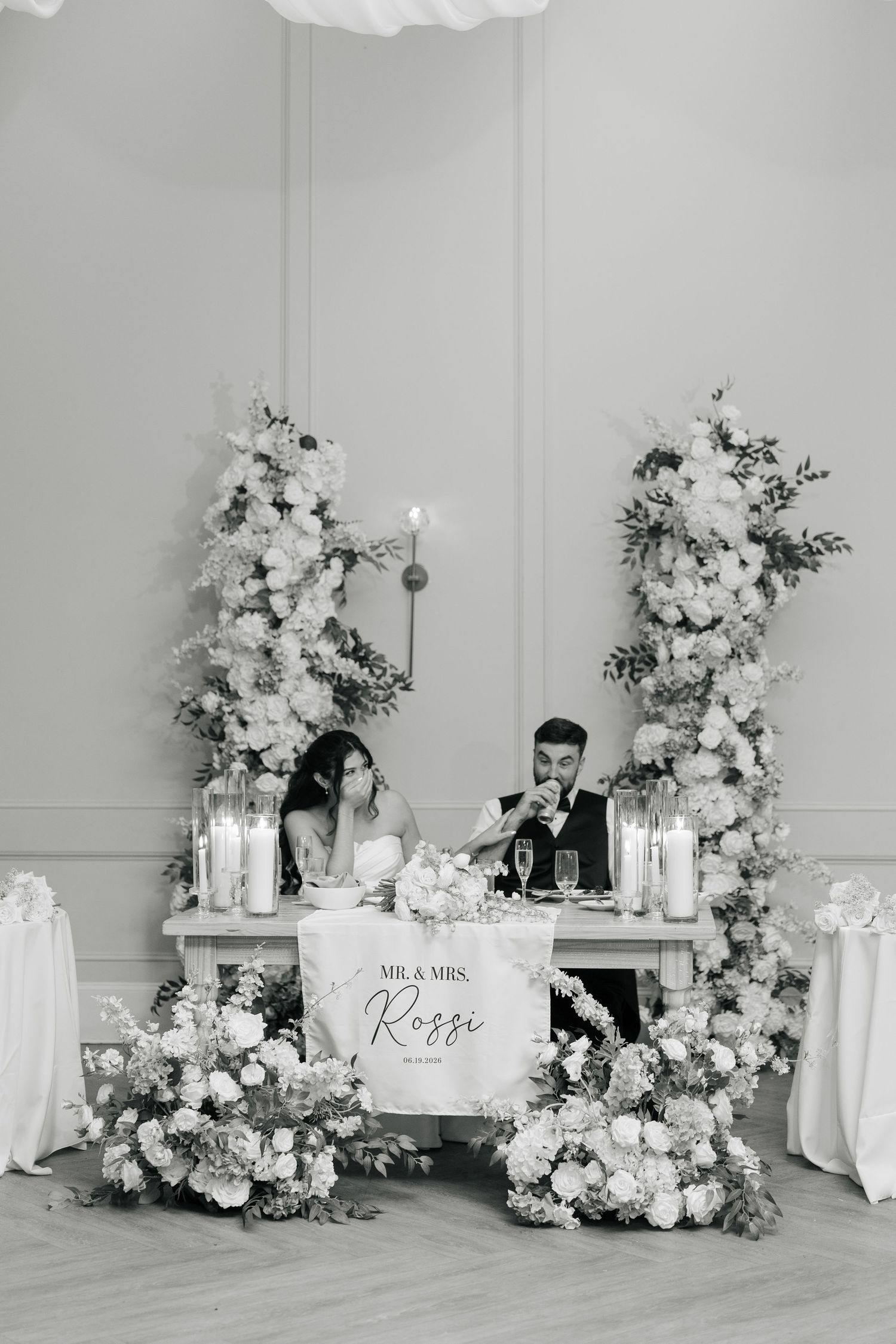 Black and white photo of Mr. & Mrs. Rossi at their floral-adorned sweetheart table with candles.