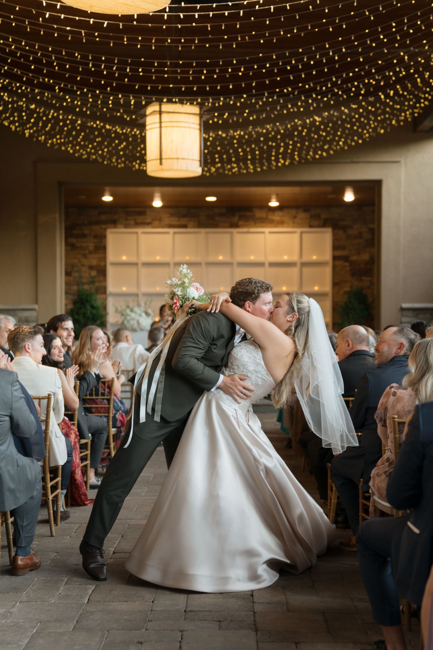 Bride and groom share a romantic dip kiss at their indoor wedding ceremony under twinkling fairy lights.