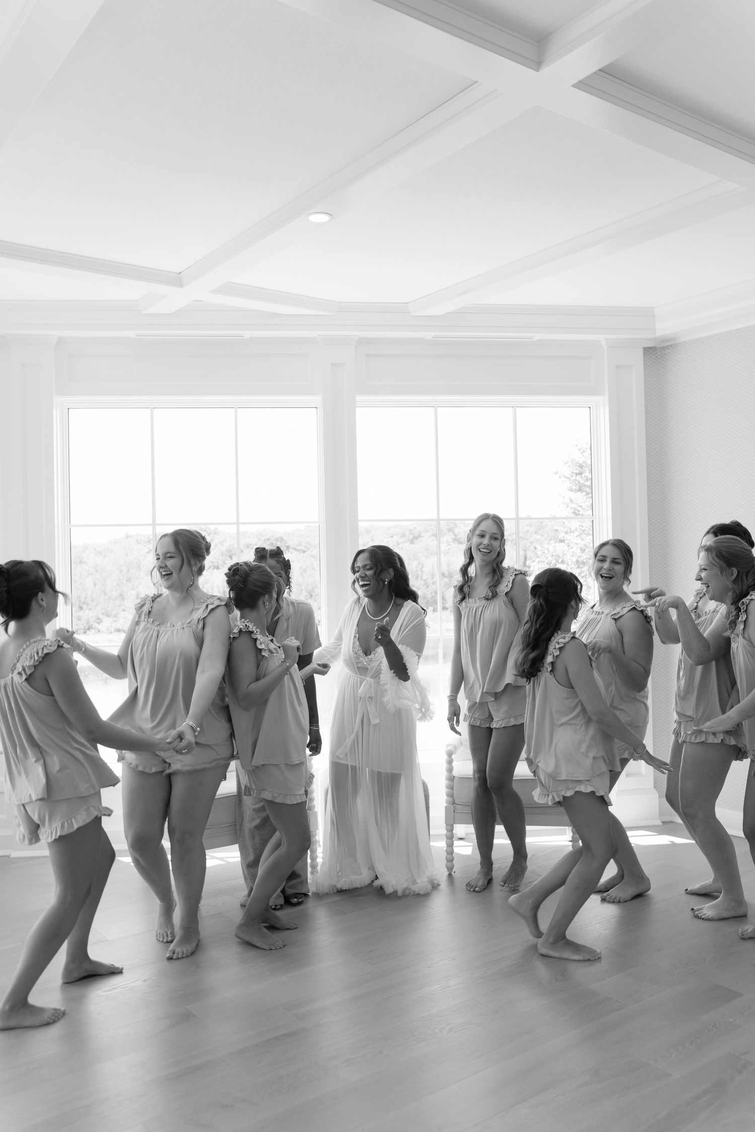 Bridesmaids in matching robes laugh and dance together in a bright white room during wedding morning preparations.
