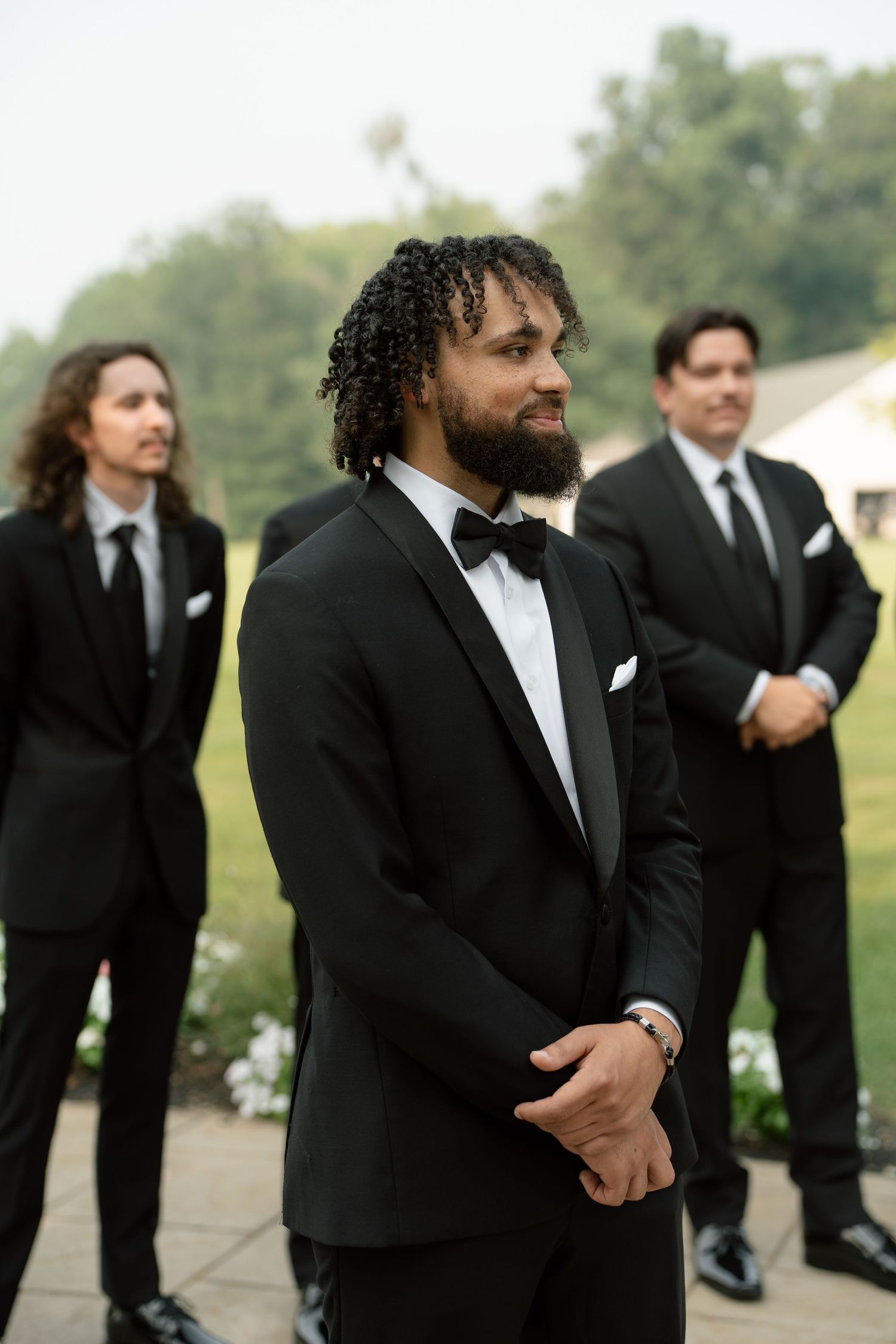 Bearded groom in black tuxedo smiles during outdoor wedding ceremony with groomsmen behind him.