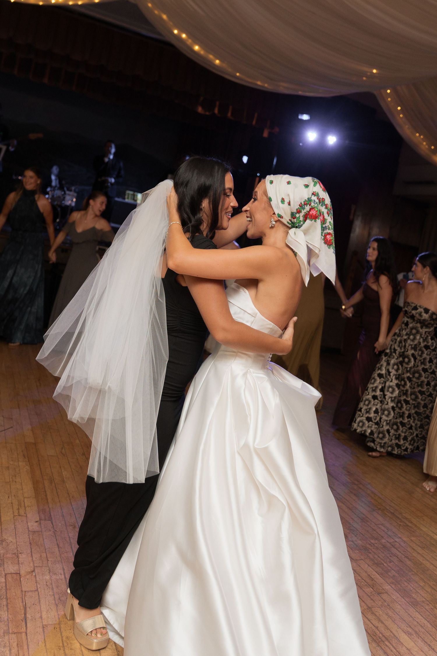 Bride in strapless white gown dancing and embracing bridesmaid wearing floral headscarf at lively wedding reception.
