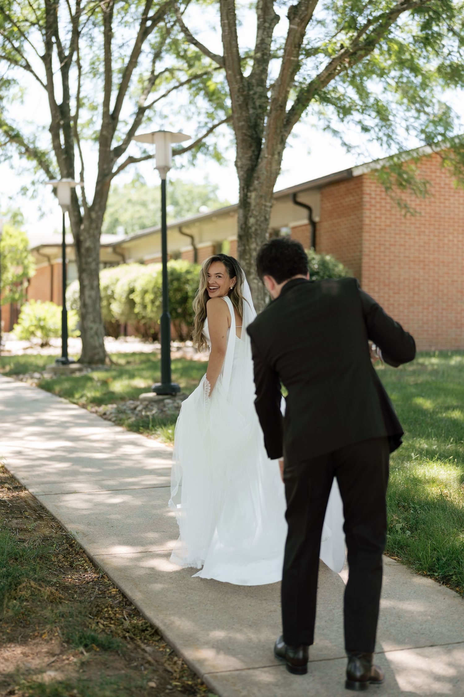 Bride in backless white gown and veil smiling as groom approaches on outdoor walkway at wedding.