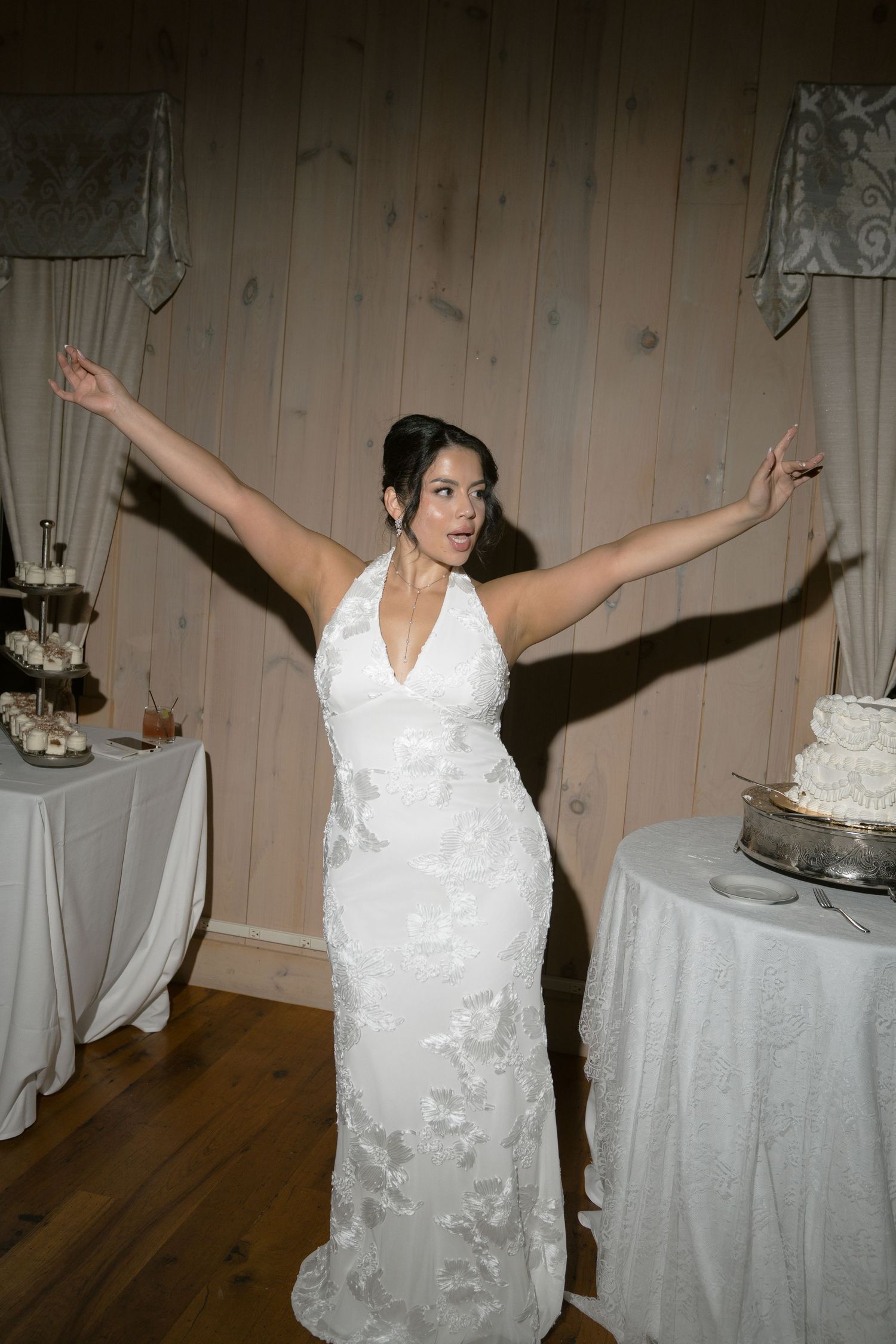 Bride in white halter wedding gown with floral appliques stands with arms outstretched near wedding cake.