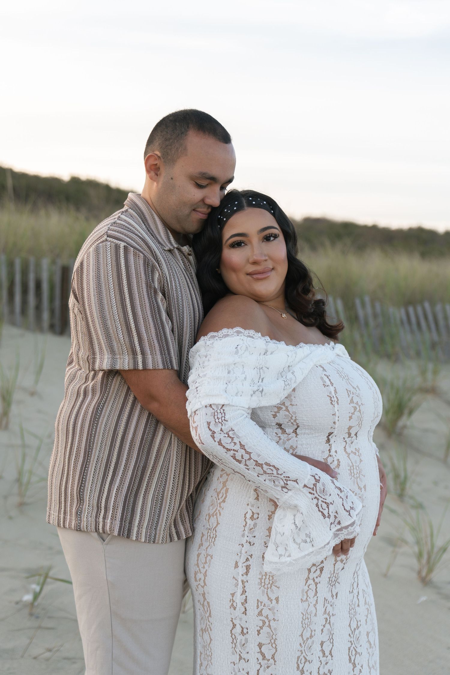 Pregnant woman in white lace off-shoulder gown with partner on sandy beach at dusk.
