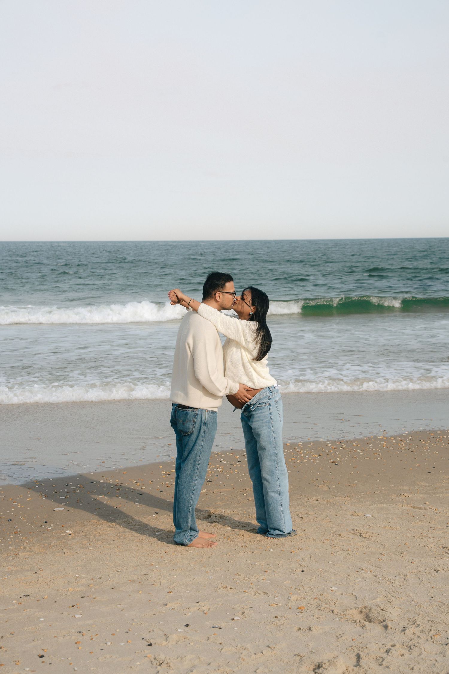 Couple in matching cream sweaters and jeans embrace on sunny beach.