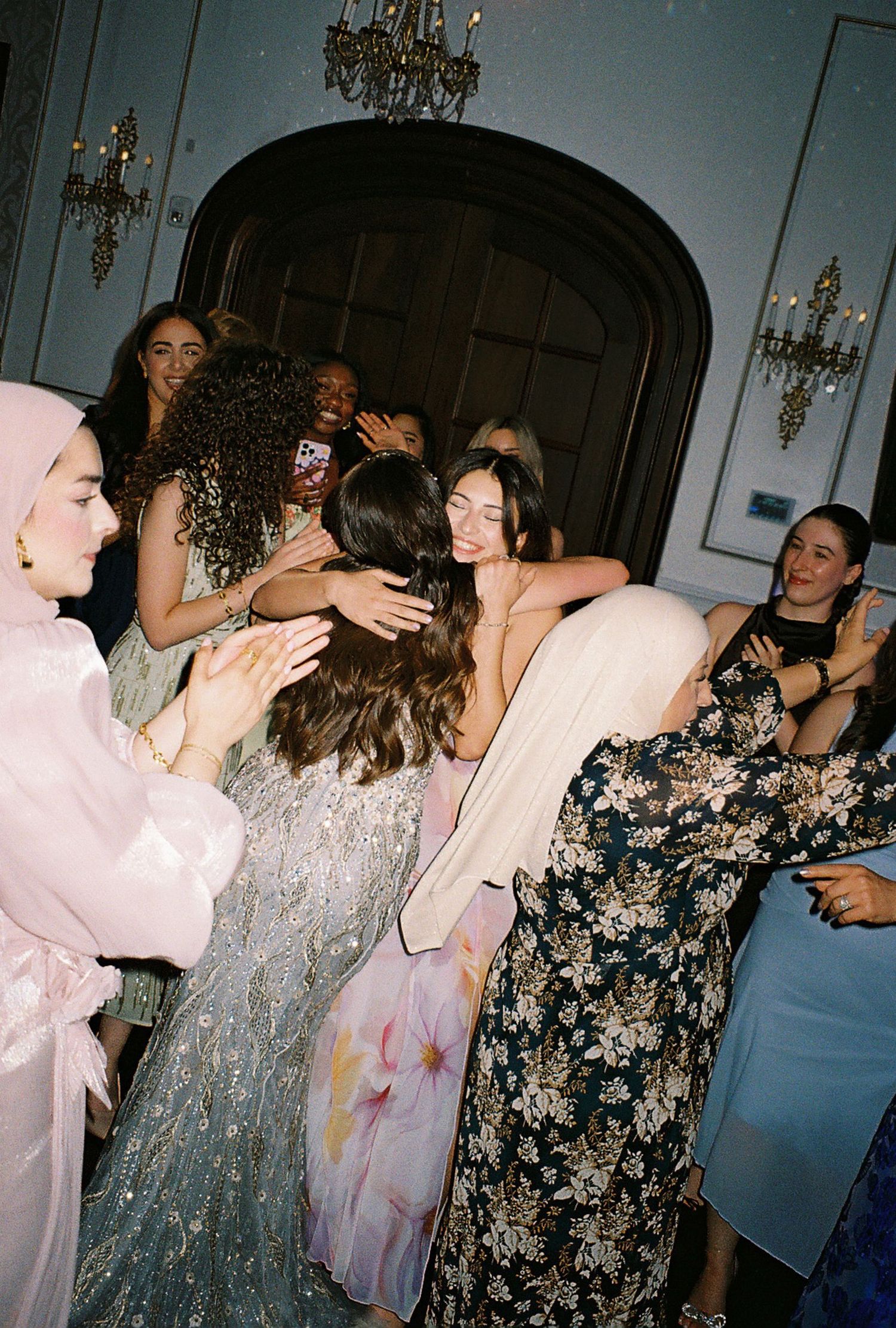 Party scene with people dancing and celebrating in formal attire under crystal chandeliers.