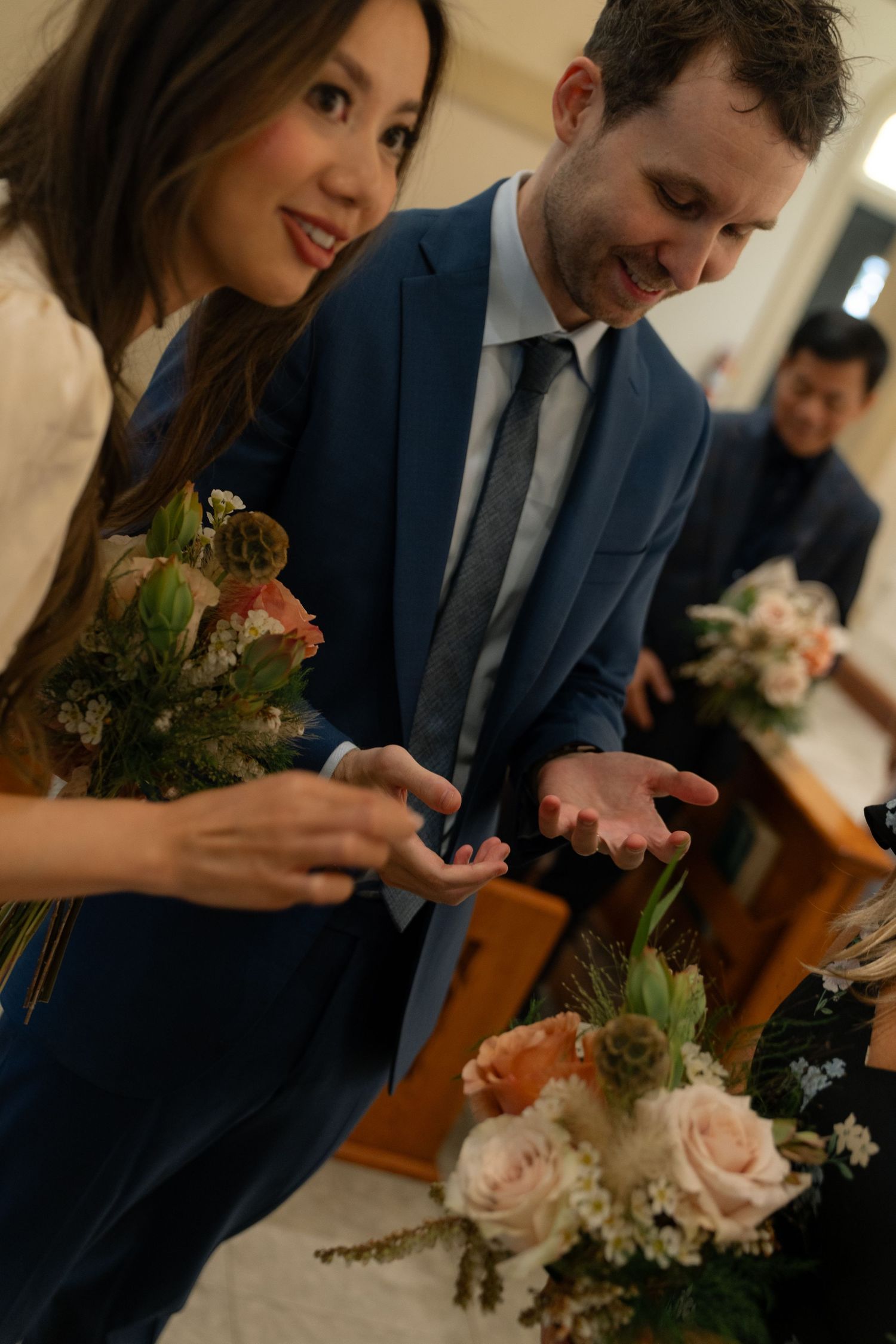Wedding party members laugh while arranging delicate pink and white floral bouquets at an indoor venue.