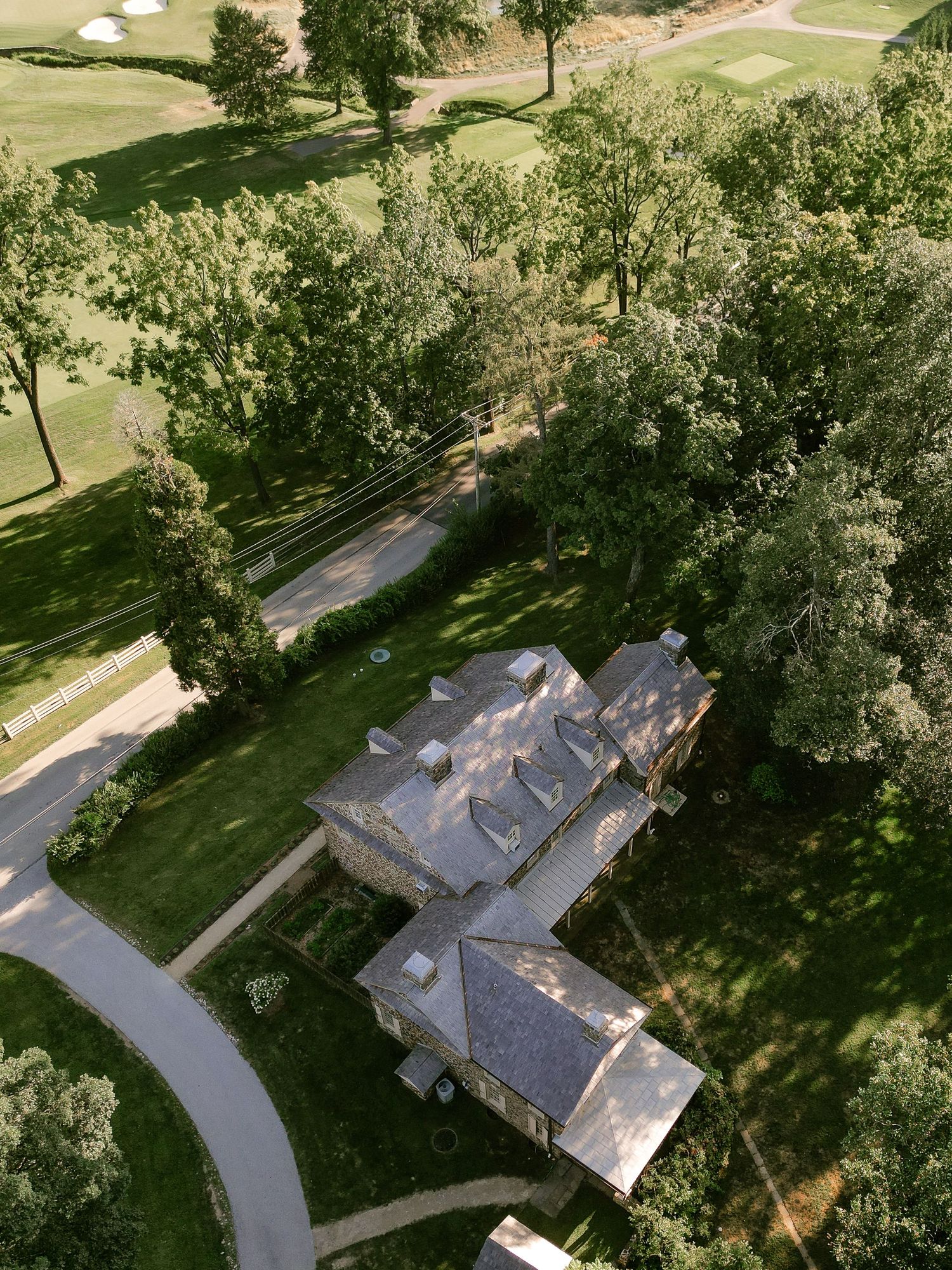 Aerial drone view of a large stone house with grey roof surrounded by trees and winding driveway.