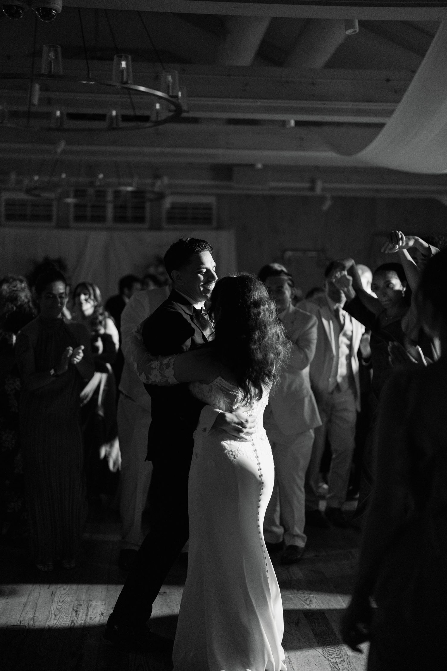 Black and white photo of bride and groom sharing first dance surrounded by guests on wedding reception dance floor.