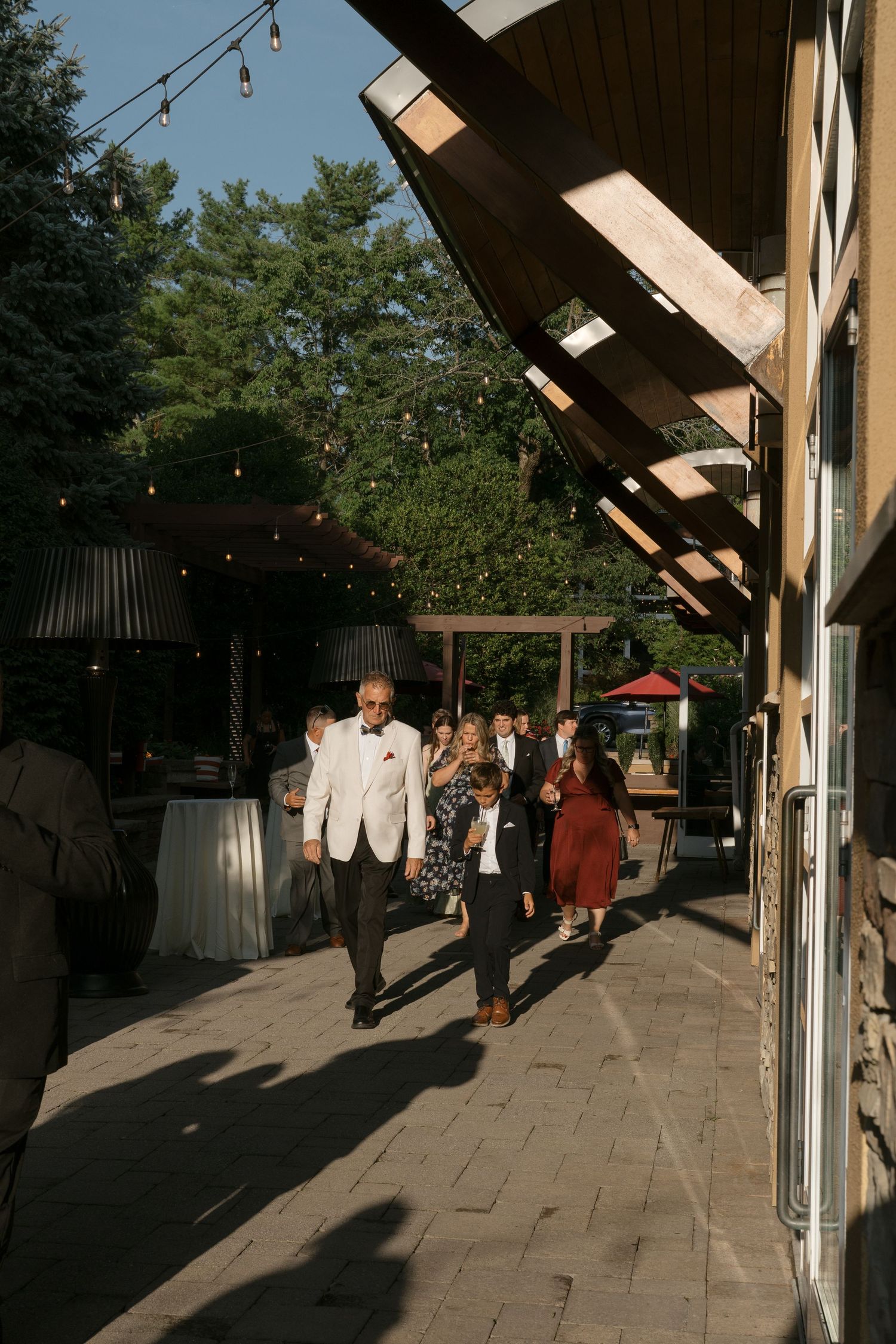 Guests in formal attire walk along a patio with string lights at an outdoor evening wedding venue.