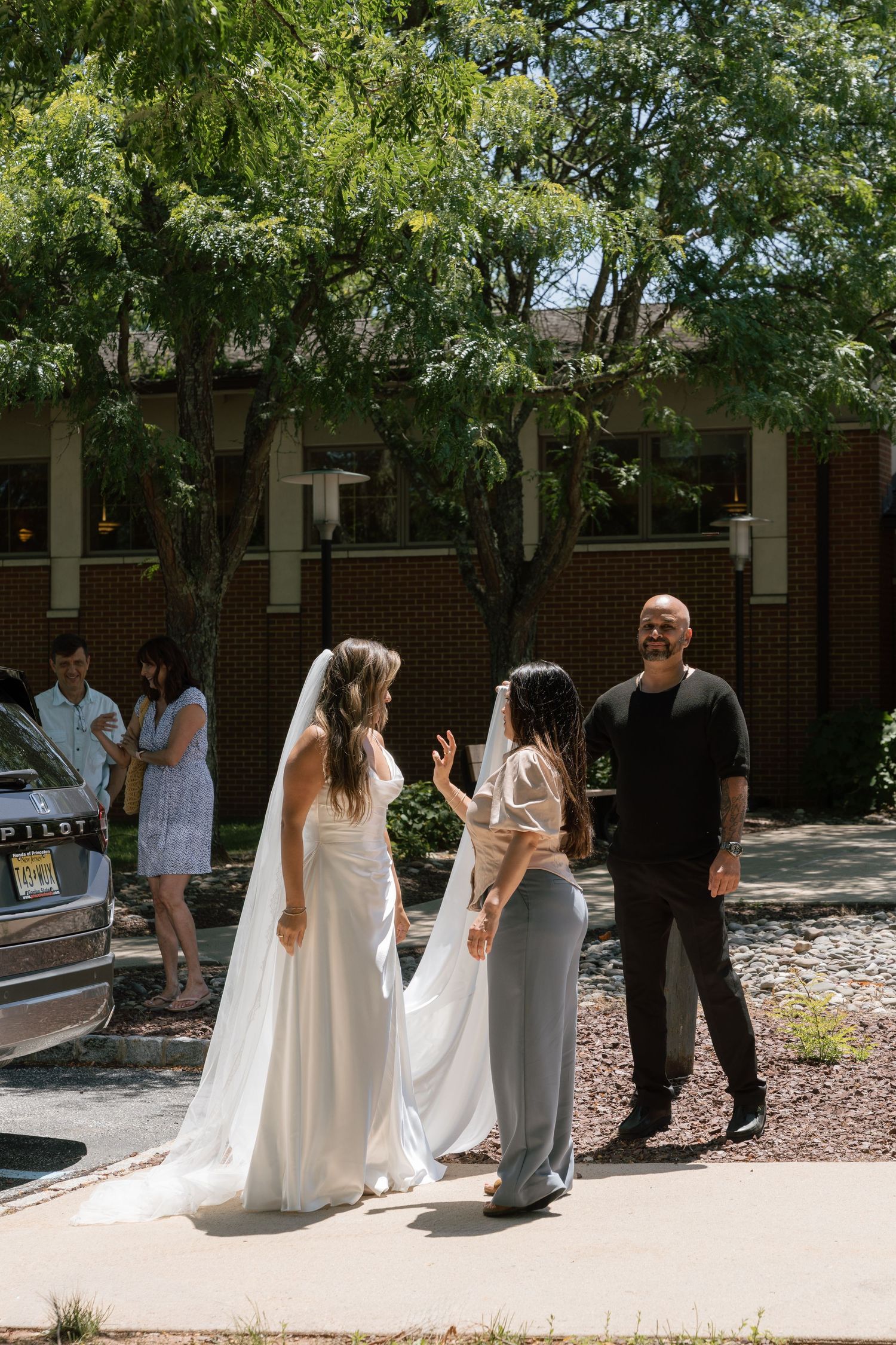 Bride in white gown and veil speaking with wedding guests outside brick building on sunny day.