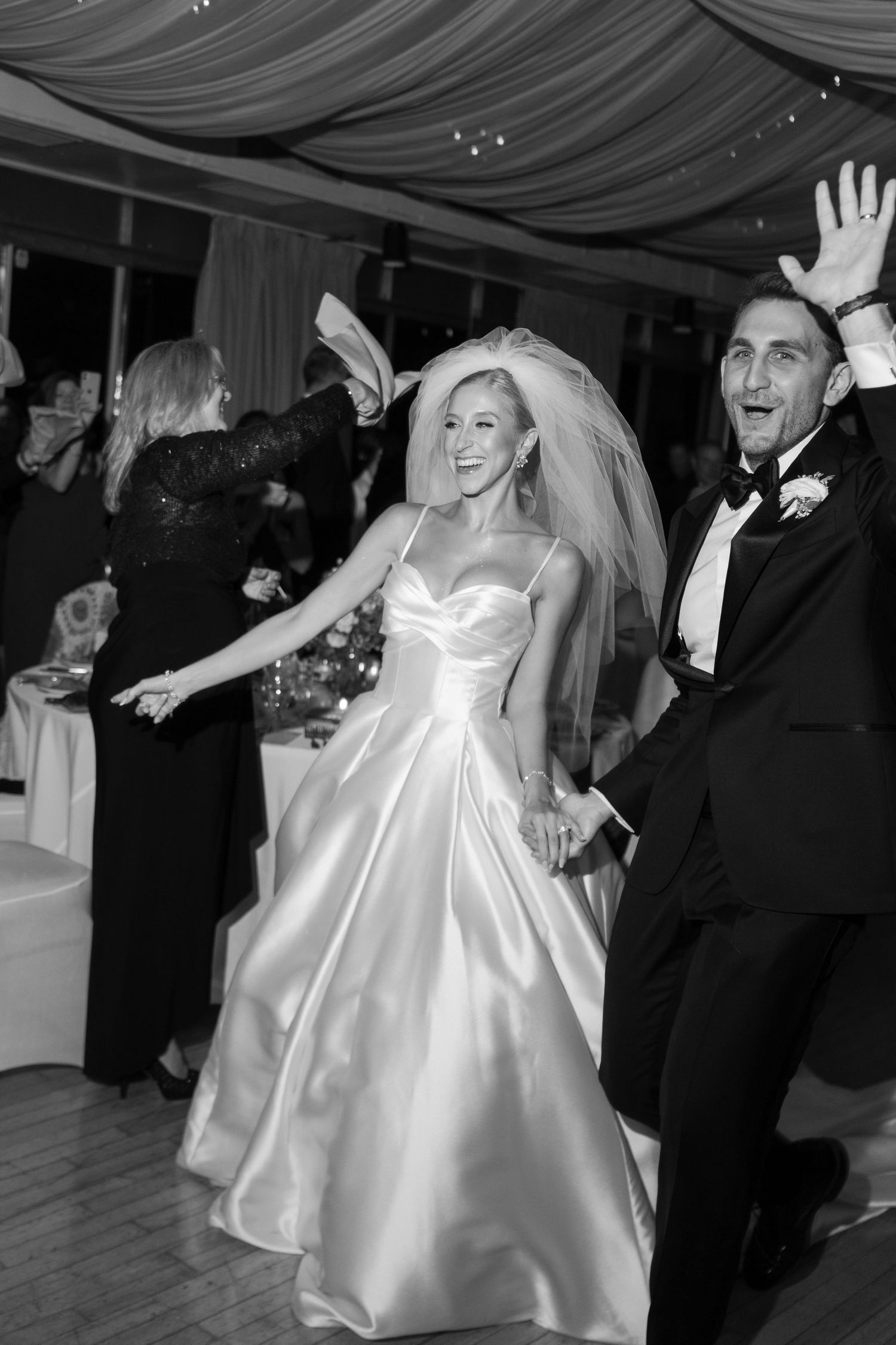 Joyful bride and groom dance energetically at wedding reception in black and white photo.