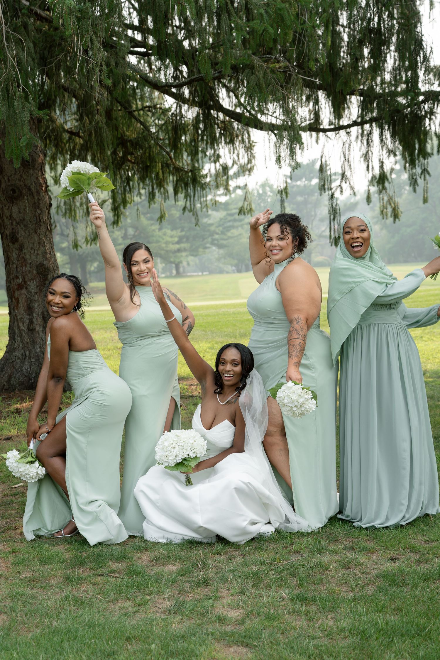 Bride in white gown poses with four bridesmaids in sage green dresses under a tree outdoors.